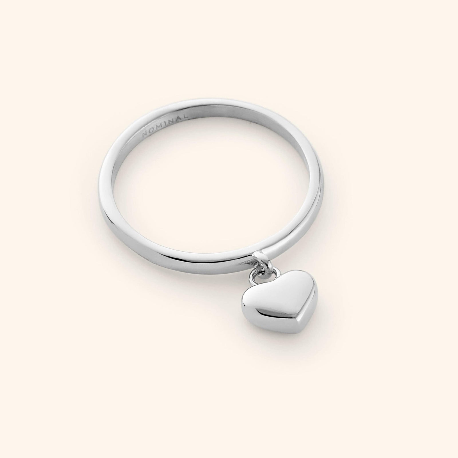 Close-up of a polished silver ring with a smooth band, featuring a small 3D heart charm dangling at the center.
