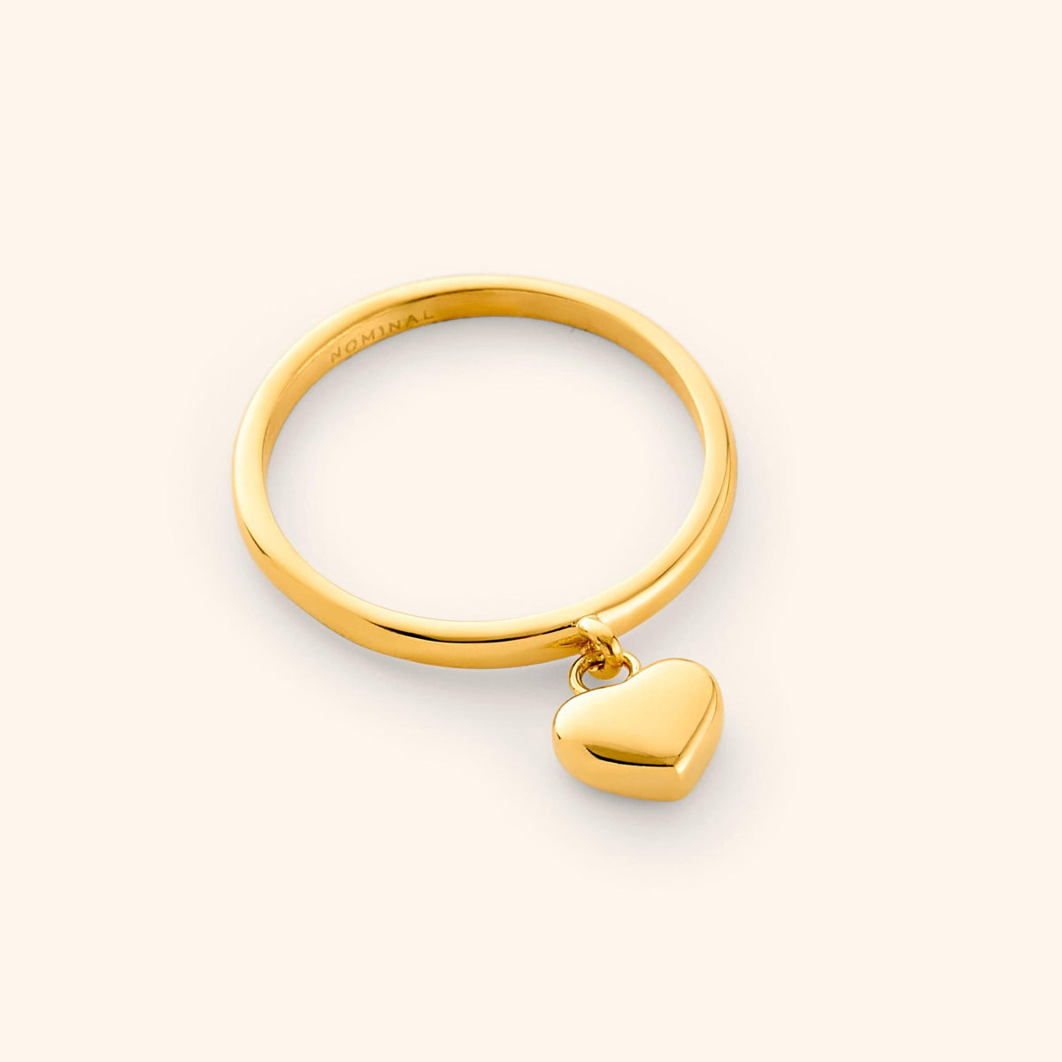 Close-up of a polished gold ring with a smooth band, featuring a small 3D heart charm dangling at the center.