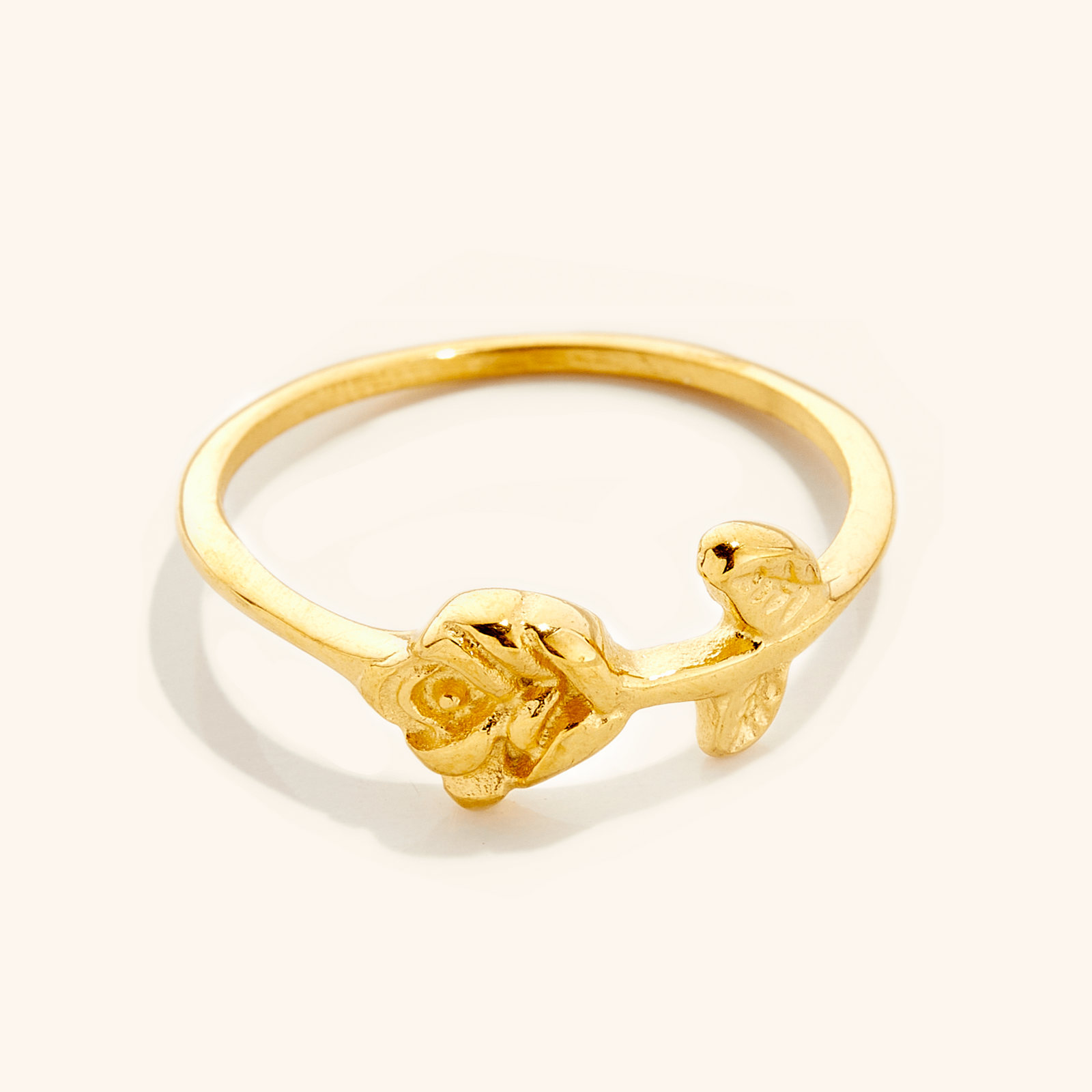 Gold ring featuring a delicate rose design with carved petals and leaves on a slim band, photographed against a neutral background.