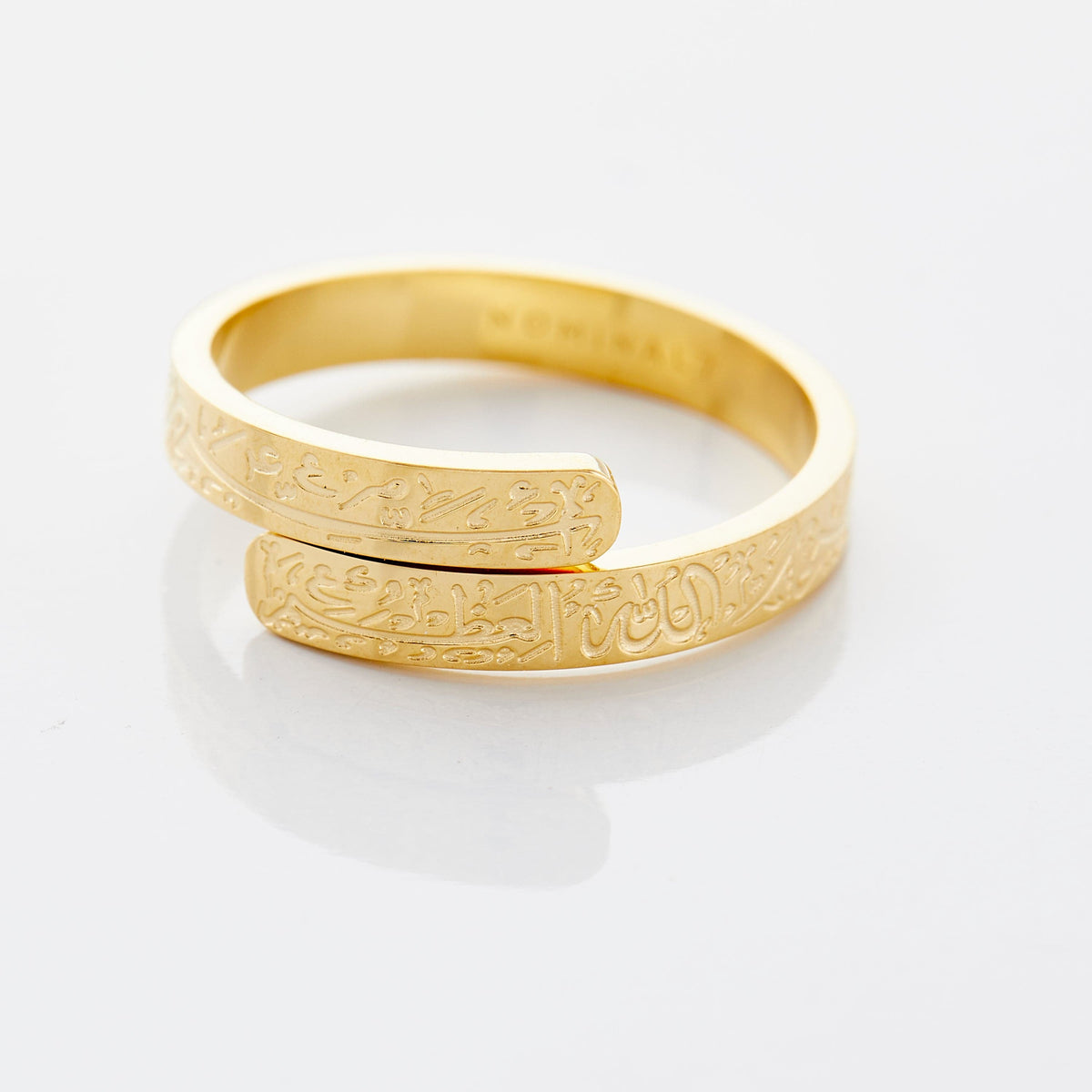 Close-up of a gold thikr wrap ring engraved with Arabic calligraphy wrapping around the band, photographed on a neutral background.