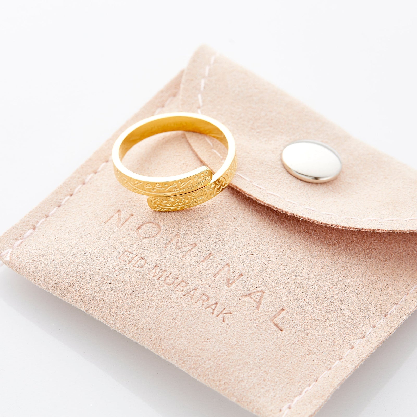 Gold wrap ring engraved with Arabic calligraphy placed on top of a tan suede Nominal pouch with “Nominal Eid Mubarak” lettering pressed
