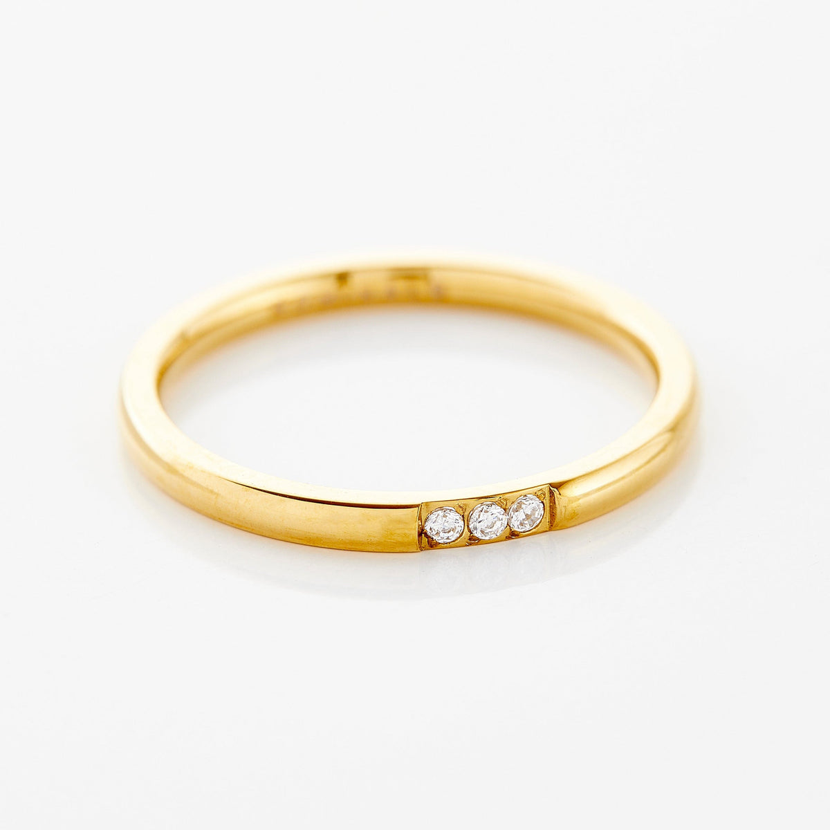 Close-up of a thin gold ring with three small clear bezel-set stones placed side by side along the band, photographed against a neutral background.