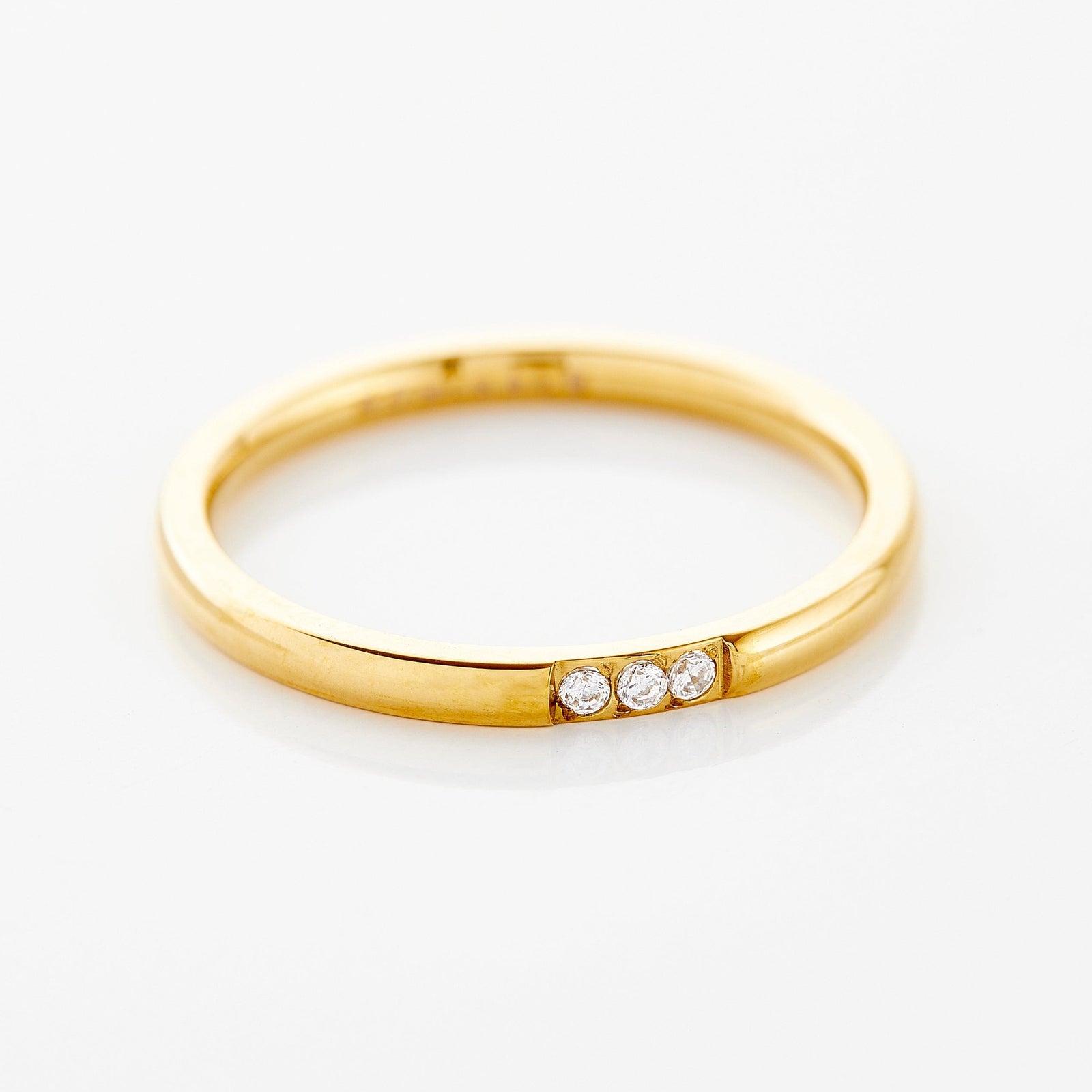 Close-up of a thin gold ring with three small clear bezel-set stones placed side by side along the band, photographed against a neutral background.