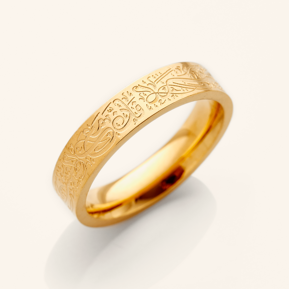 A gold ring with an intricate design etched around the outer surface. The design appears to be Arabic script meaning "With Hardship Comes Ease". The ring is displayed on a bright white background with a soft shadow underneath. 