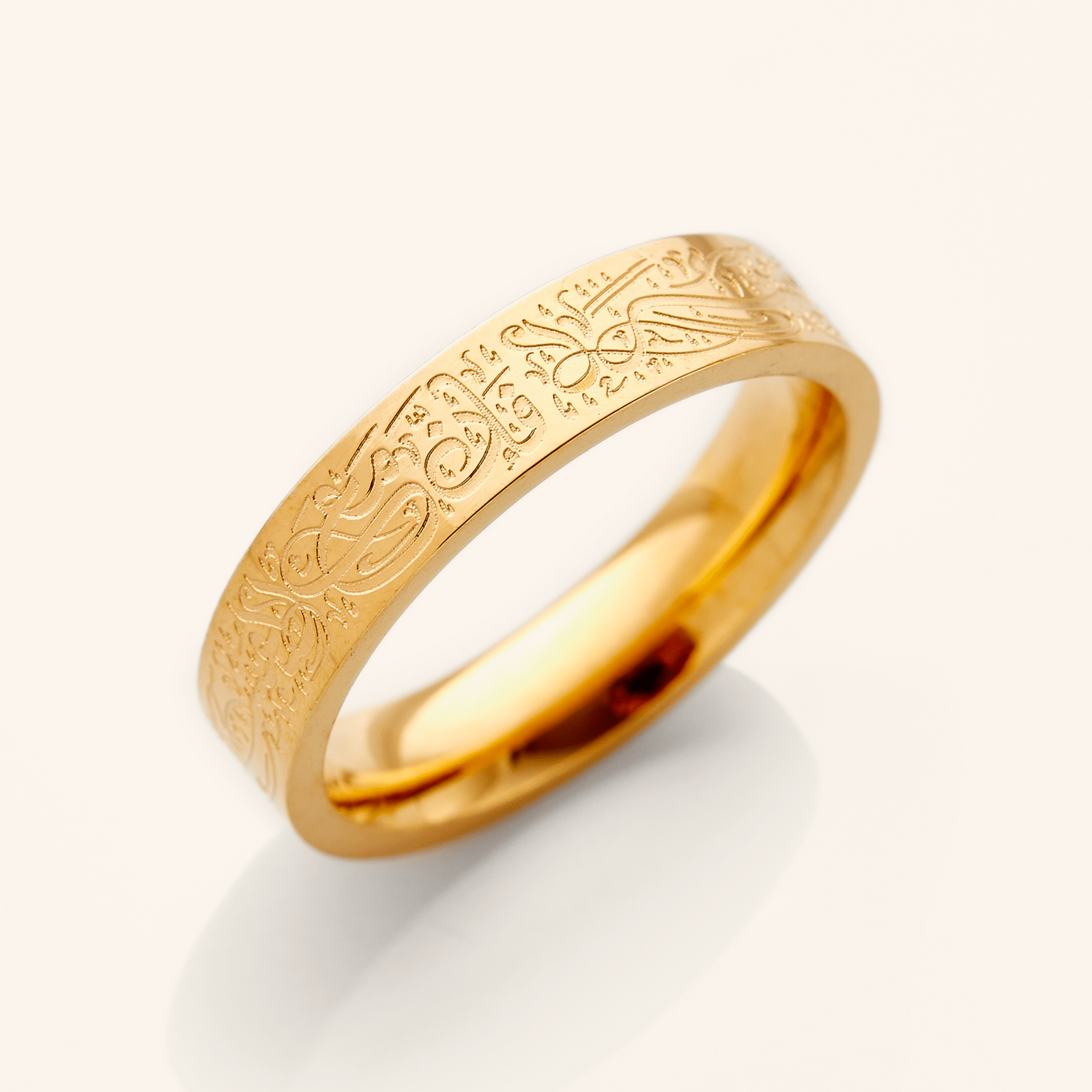 A gold ring with an intricate design etched around the outer surface. The design appears to be Arabic script meaning "With Hardship Comes Ease". The ring is displayed on a bright white background with a soft shadow underneath. 