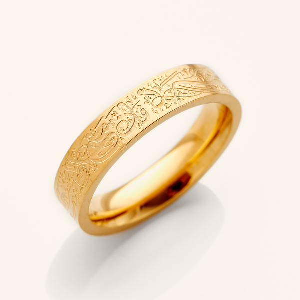 A gold ring with an intricate design etched around the outer surface. The design appears to be Arabic script meaning "With Hardship Comes Ease". The ring is displayed on a bright white background with a soft shadow underneath. 