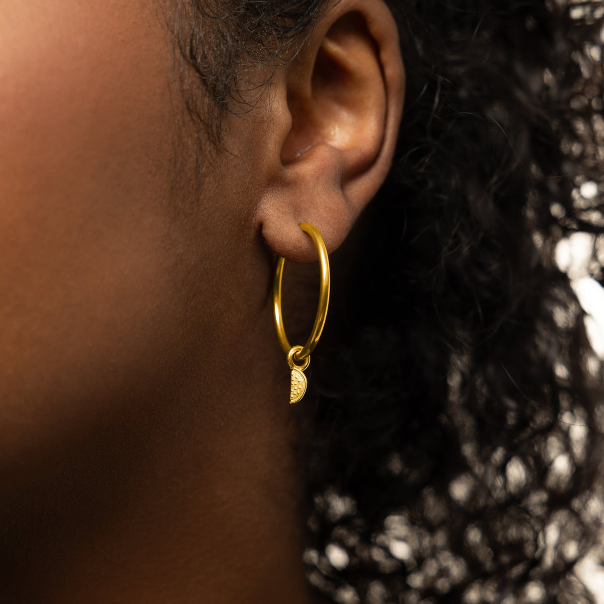 Close up of the gold Watermelon Earring Hoop Charm hanging on a gold hoop earring as worn by the model on their left ear.