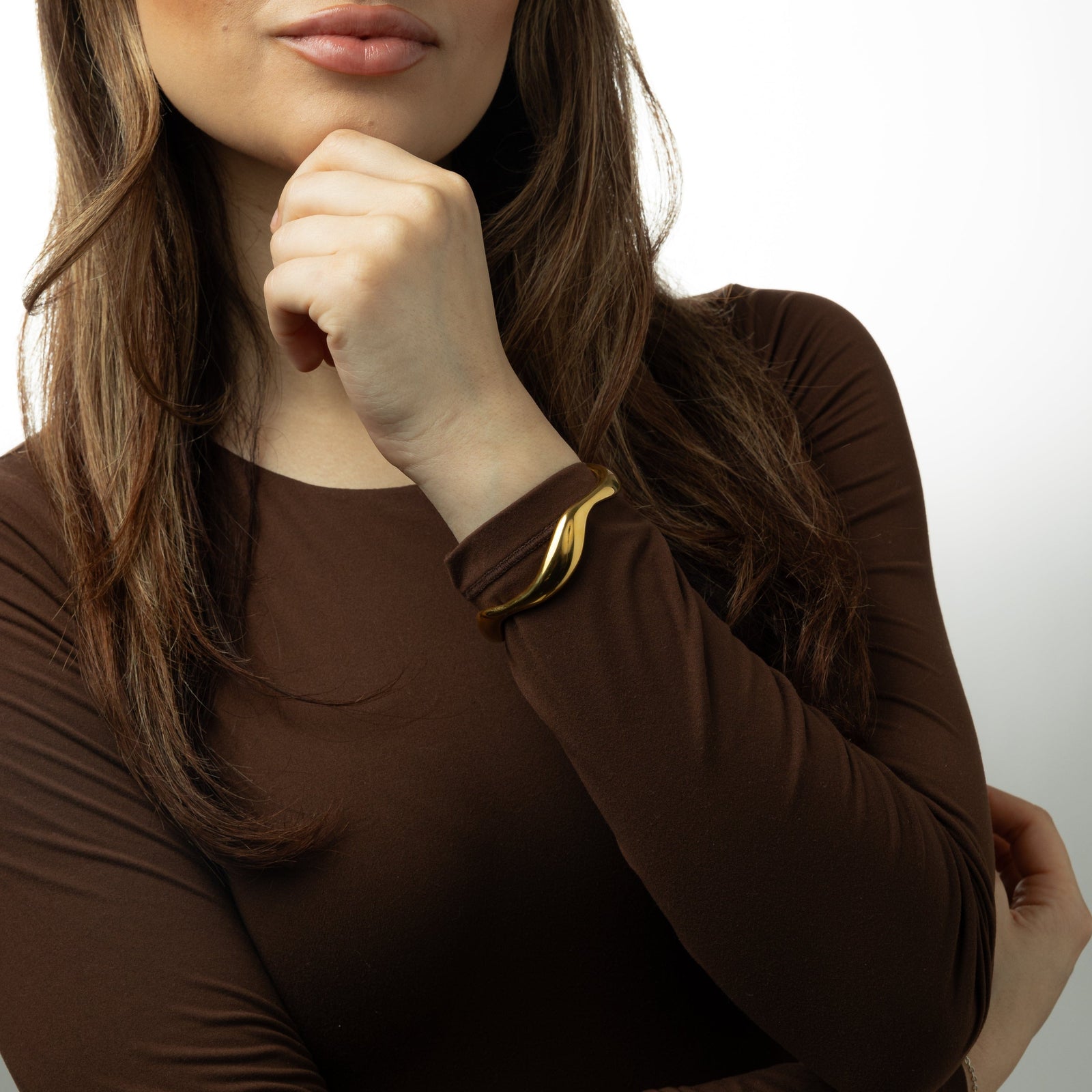 Photo of the model touching their chin with their left fingers. The model is wearing a brown long sleeves shirt and the gold Wave bangle on their left wrist.