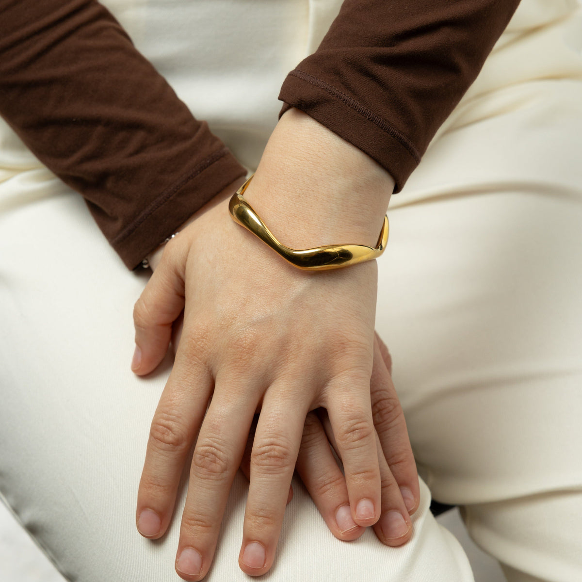 Photo of the model's left hand on top of their right hand. The model is wearing the gold Wave Bangle on their left wrist.
