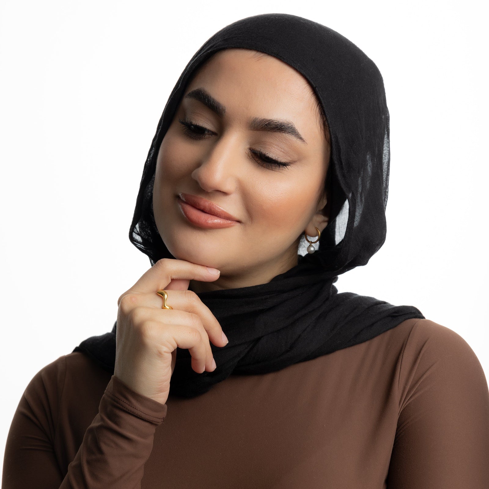 Woman in a black hijab smiling with hand near chin, wearing a gold wave ring.