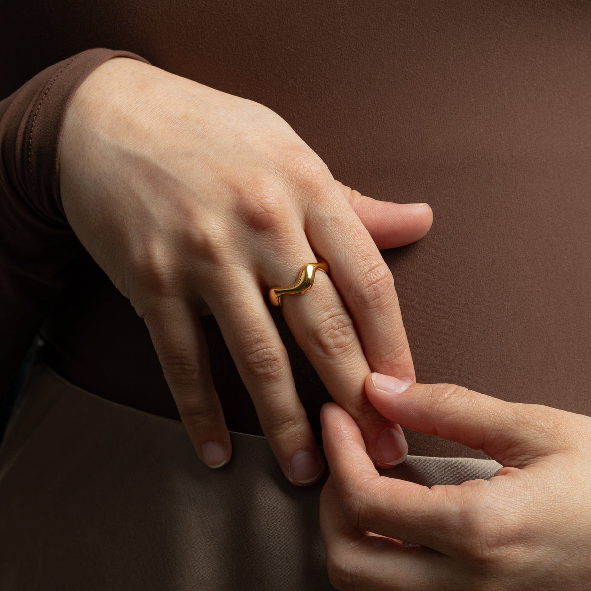 Hand wearing a gold wave ring with smooth curves, styled with a brown long-sleeve top.