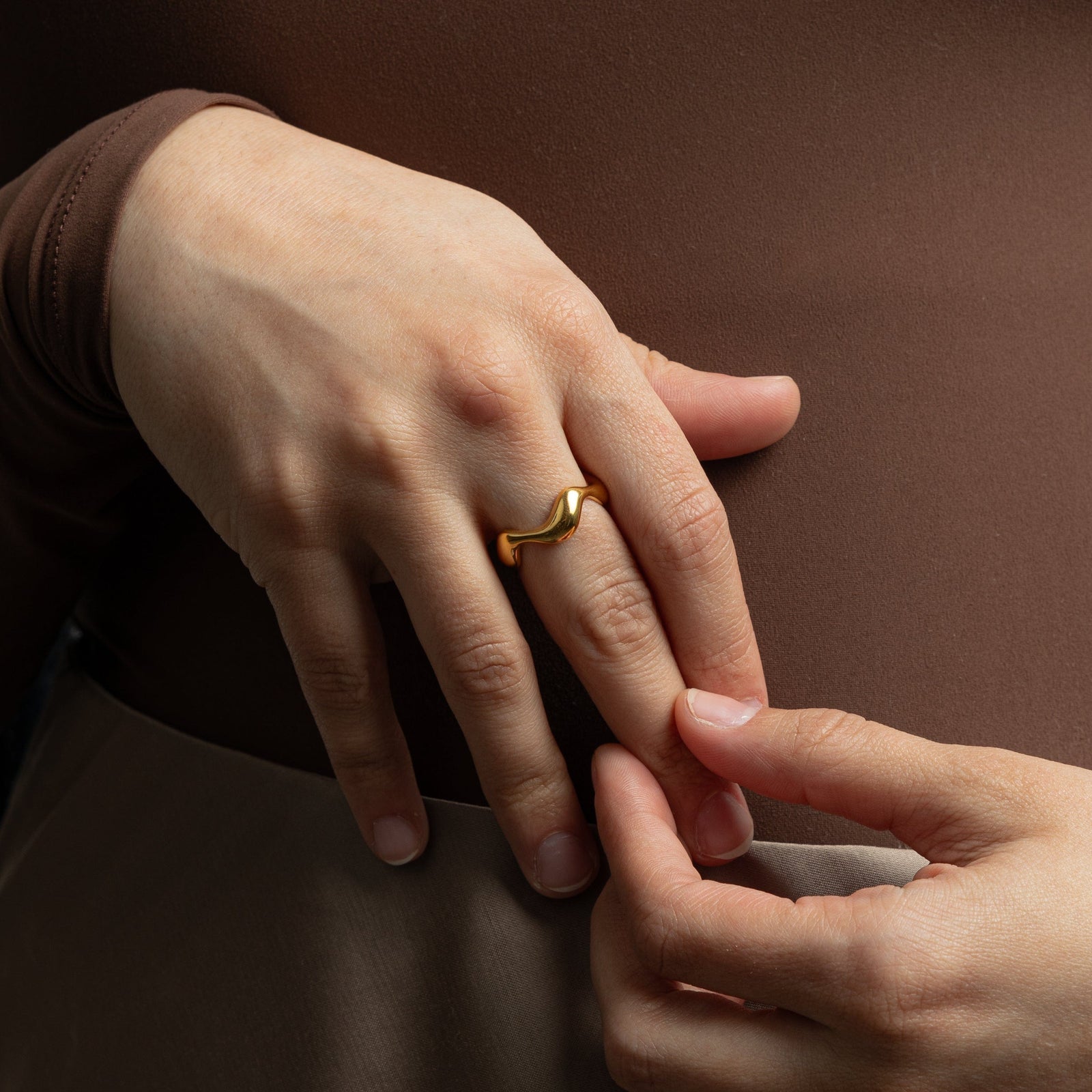 Hand wearing a gold wave ring with smooth curves, styled with a brown long-sleeve top.