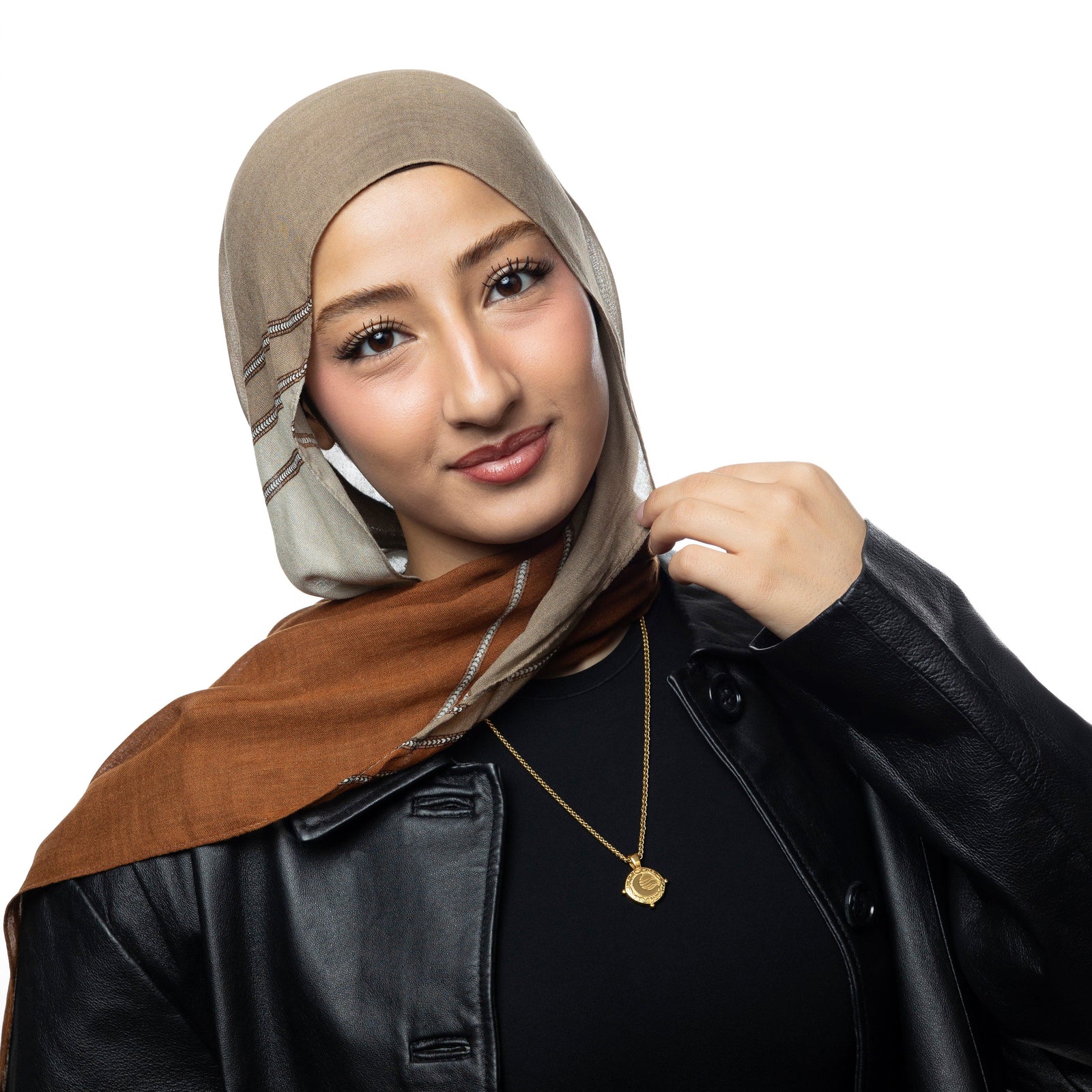 Woman wearing a gold what you seek is seeking you necklace, featuring a crescent moon detail. Styled with a beige and rust brown hijab, black top, and leather jacket.