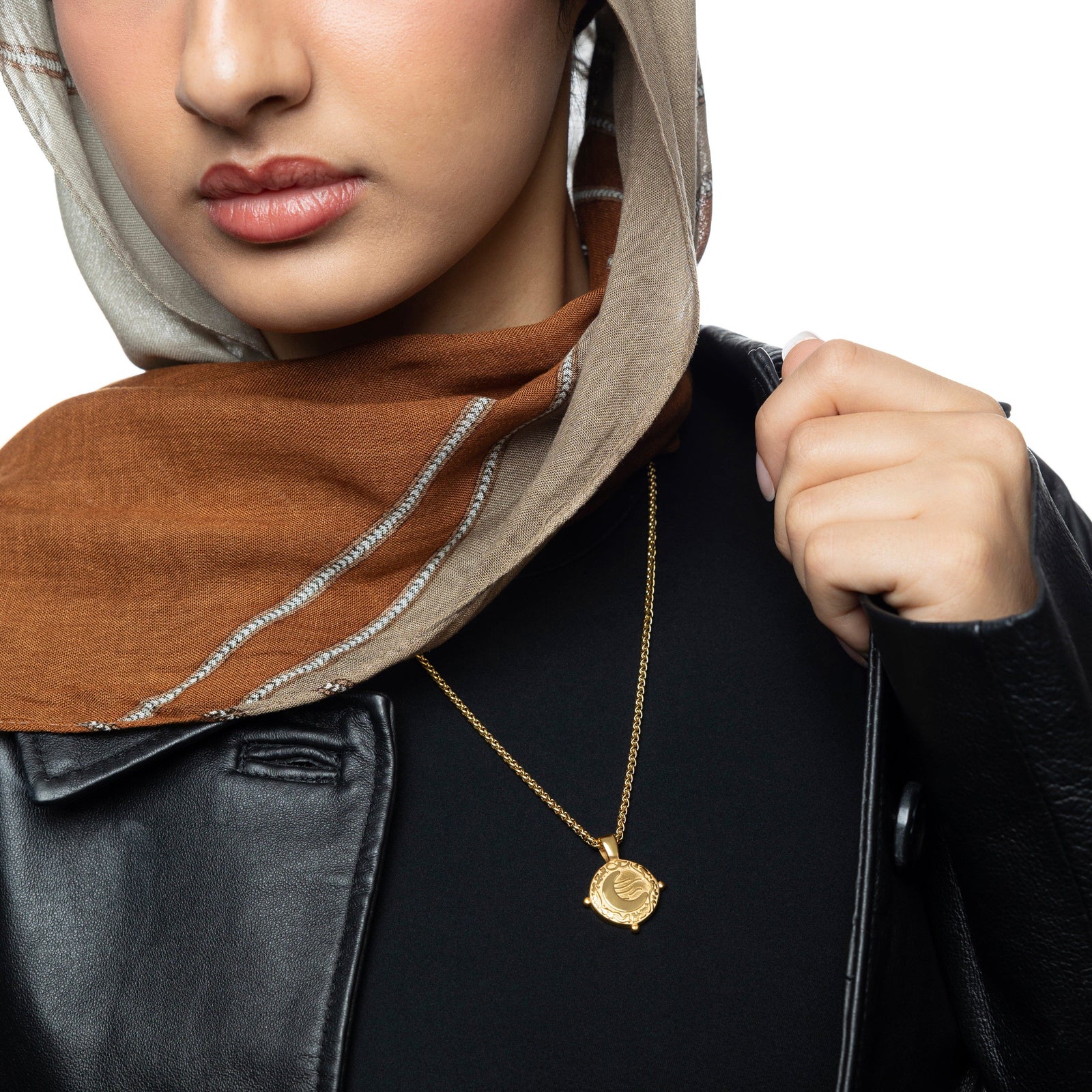 Woman wearing a gold what you seek is seeking you necklace, featuring a crescent moon Styled with a beige and rust brown hijab, black top, and leather jacket.