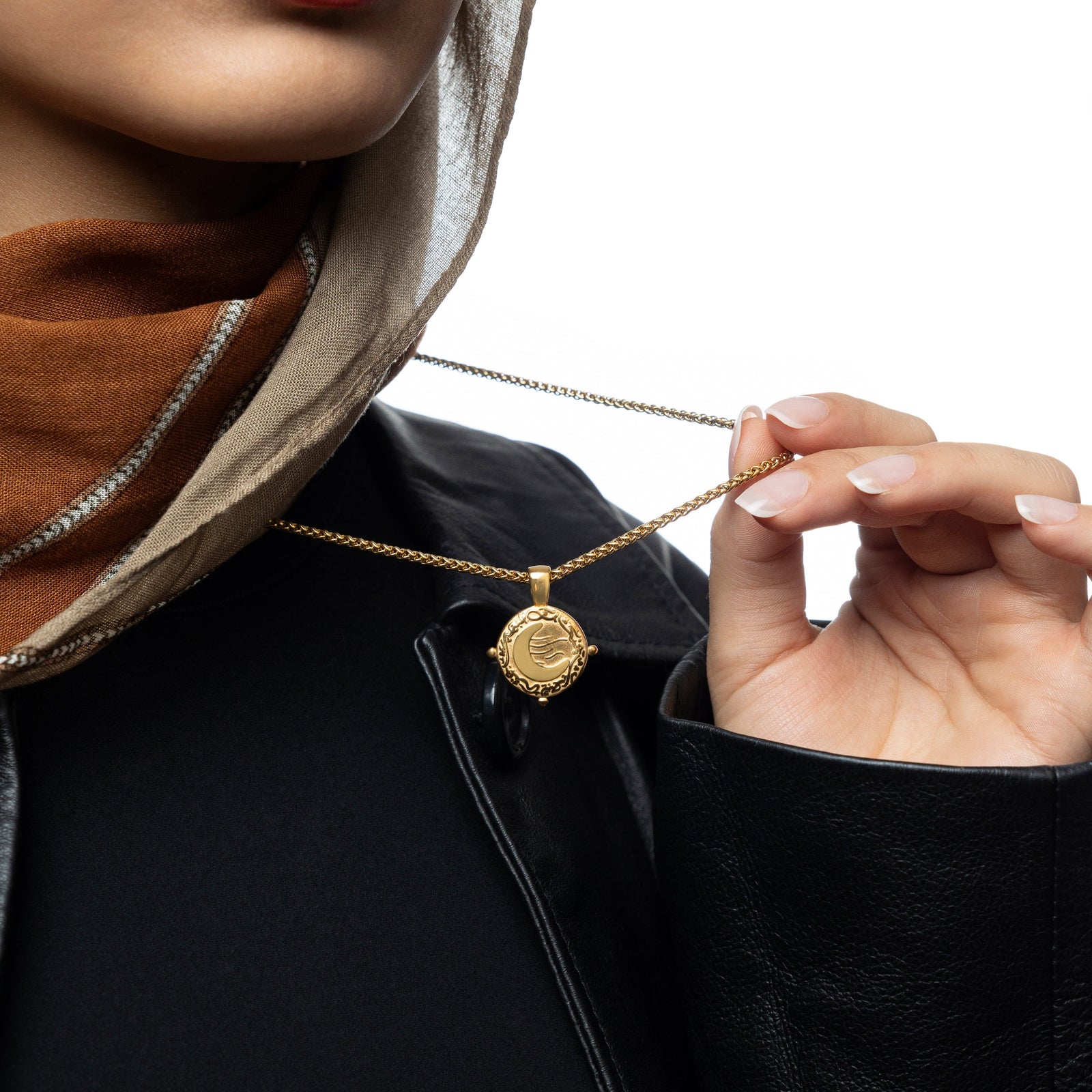 Close-up of woman holding a gold what you seek is seeking you necklace, styled over a black outfit and rust-toned scarf.