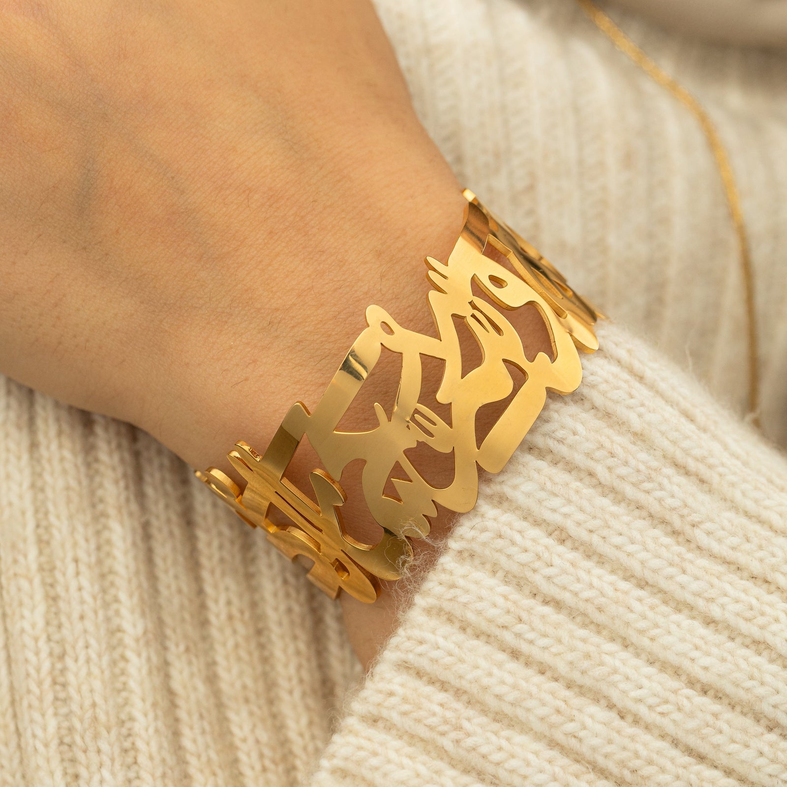 Close-up of the ‘What Makes You Different Makes You Beautiful’ Calligraphy Cuff in gold on a wrist, styled with a cream knit sweater.