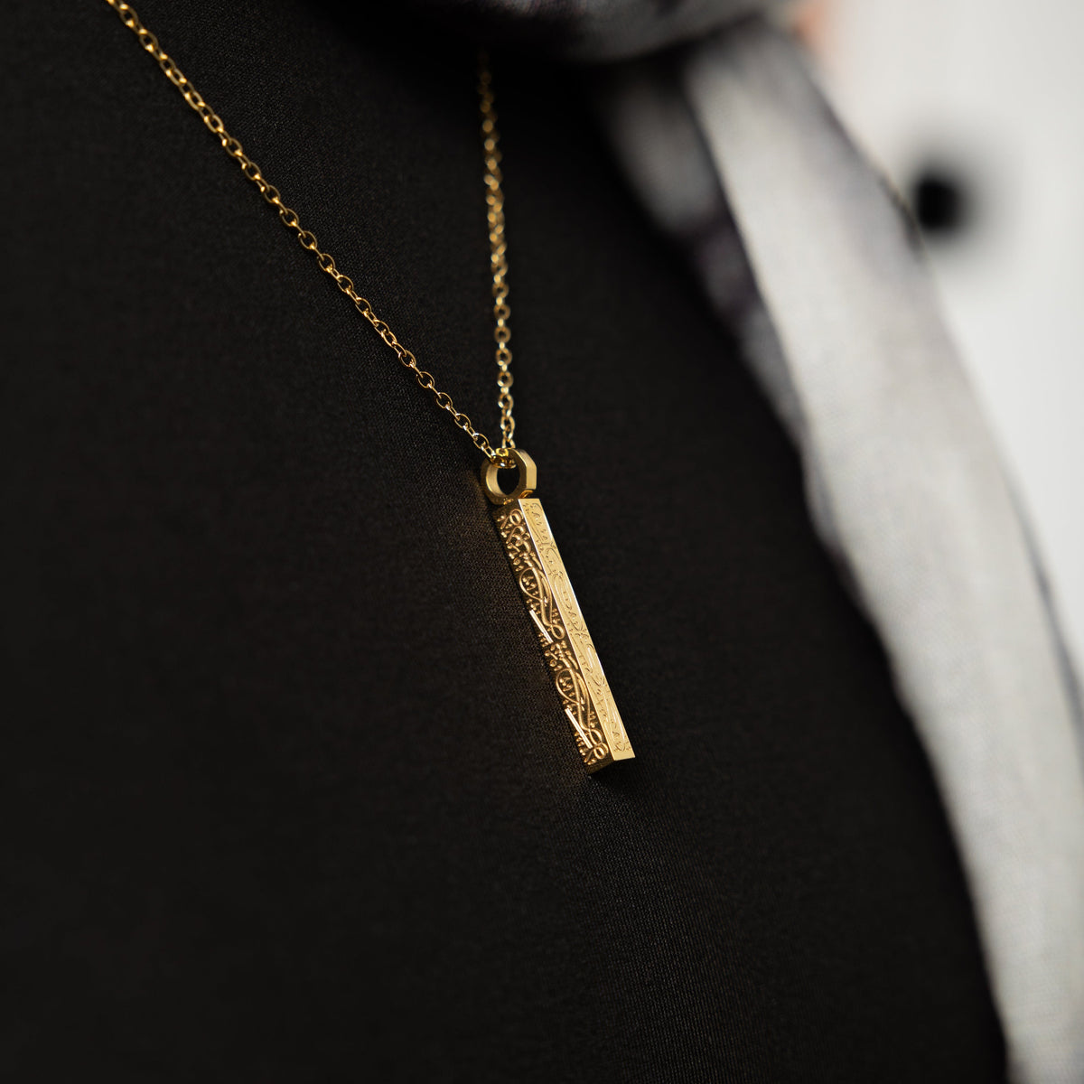 Close up of the gold pendant of the "With Hardship Comes Ease" Bar against the dark fabric of the model's shirt.