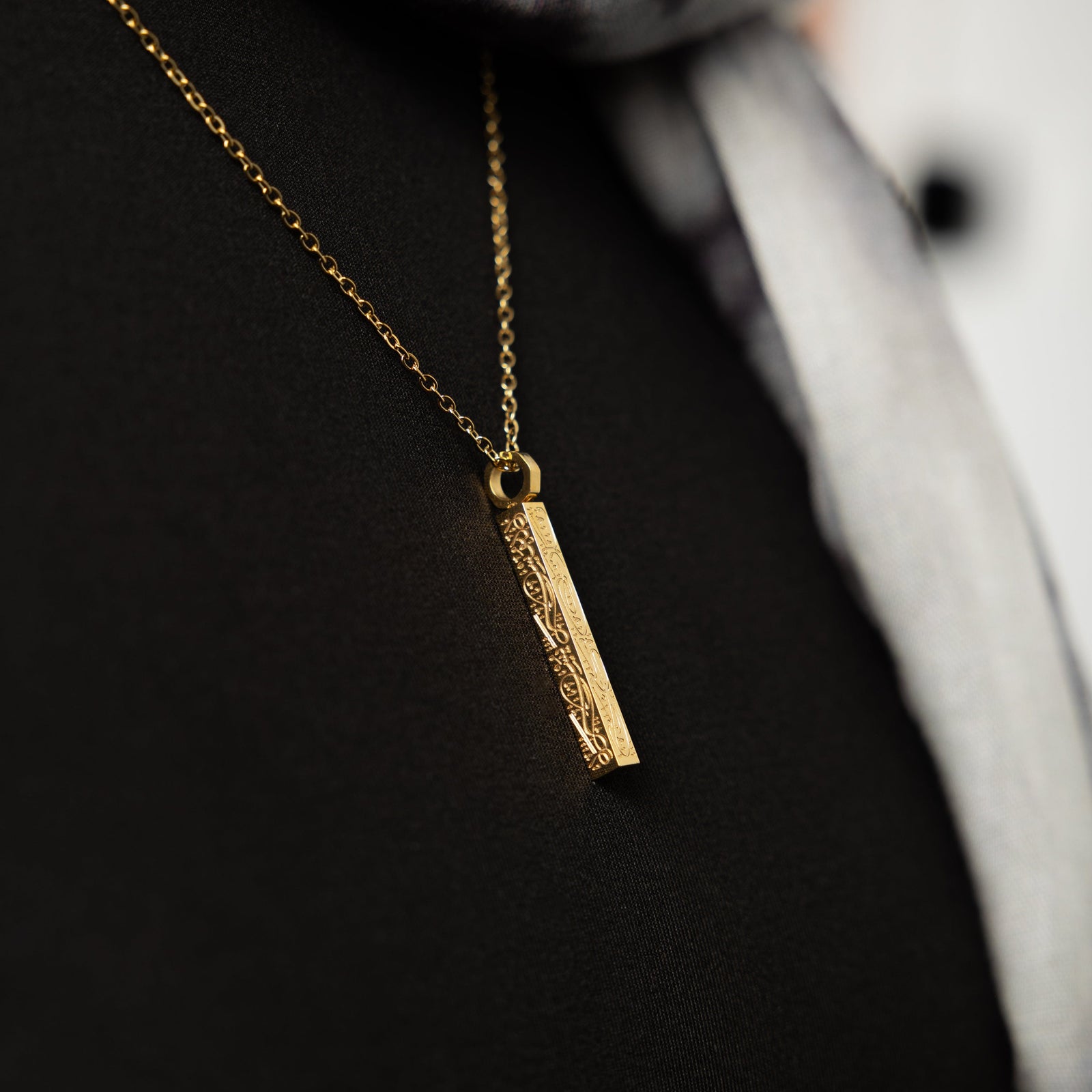 Close up of the gold pendant of the "With Hardship Comes Ease" Bar against the dark fabric of the model's shirt.