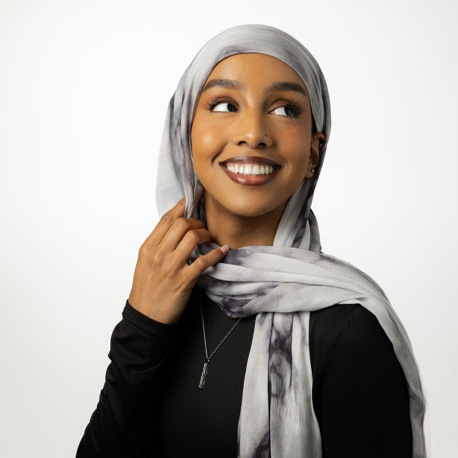 Photo of the model wearing a black long-sleeve top along with a light gray scarf that is wrapped around her head and draped over her shoulders. The scarf has soft, subtle patterns that add texture and contrast to the outfit. The combination of the dark clothing and the light scarf creates a balanced and stylish look.

The woman is also wearing delicate jewelry, including a thin necklace with a small vertical pendant that hangs at the center of her chest.