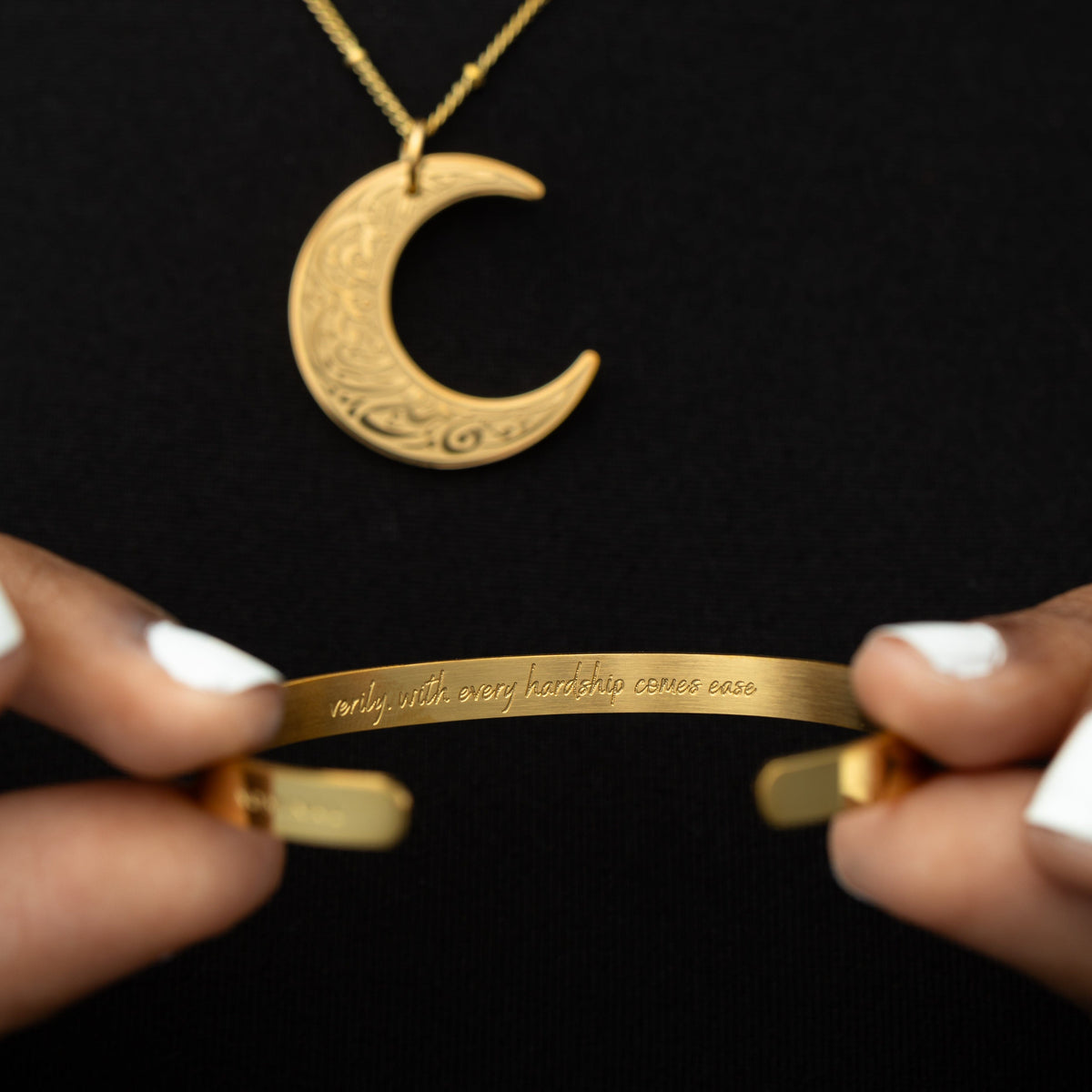 Close up of the gold crescent necklace and the gold cuff against the black fabric of the model's clothing. The model is holding the cuff to show the engraved writing, "Verily, with hardship comes ease."