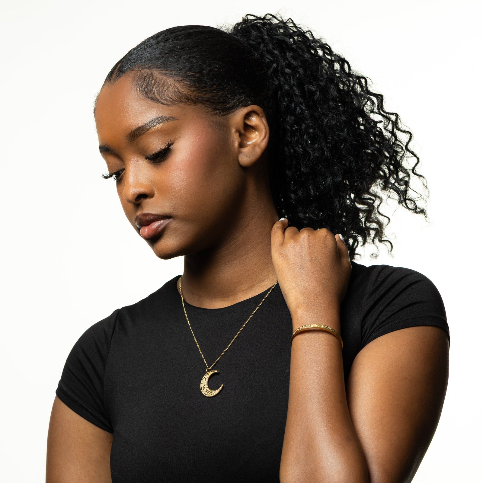Photo of the model looking down and holding the back of their neck, near their left shoulder, with their left hand. The model is wearing both pieces of the  gold "With Hardship Comes Ease" Gift Set.