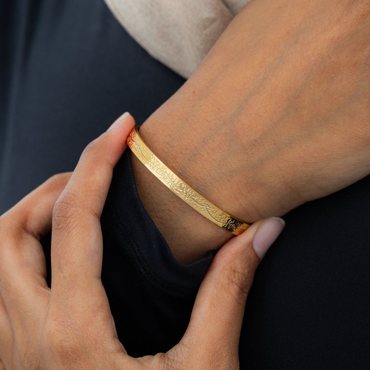A model wearing the 18K gold-plated "With Hardship Comes Ease" Cuff V2. The cuff rests gracefully on her wrist, showcasing the embossed Arabic calligraphy against her skin, symbolizing resilience and hope.