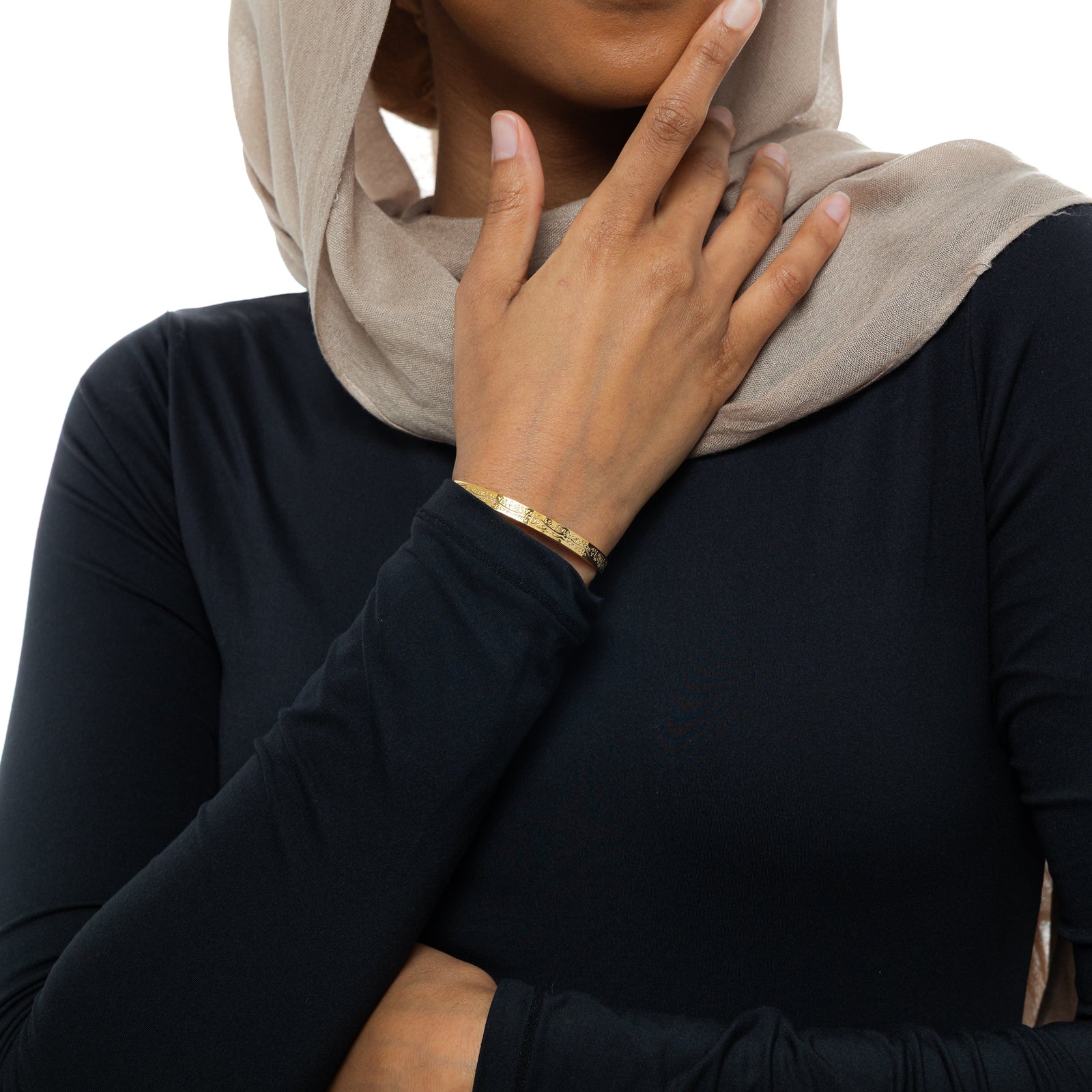 A model wearing the 18K gold-plated "With Hardship Comes Ease" Cuff V2. The cuff rests gracefully on her wrist, showcasing the embossed Arabic calligraphy against her skin, symbolizing resilience and hope.