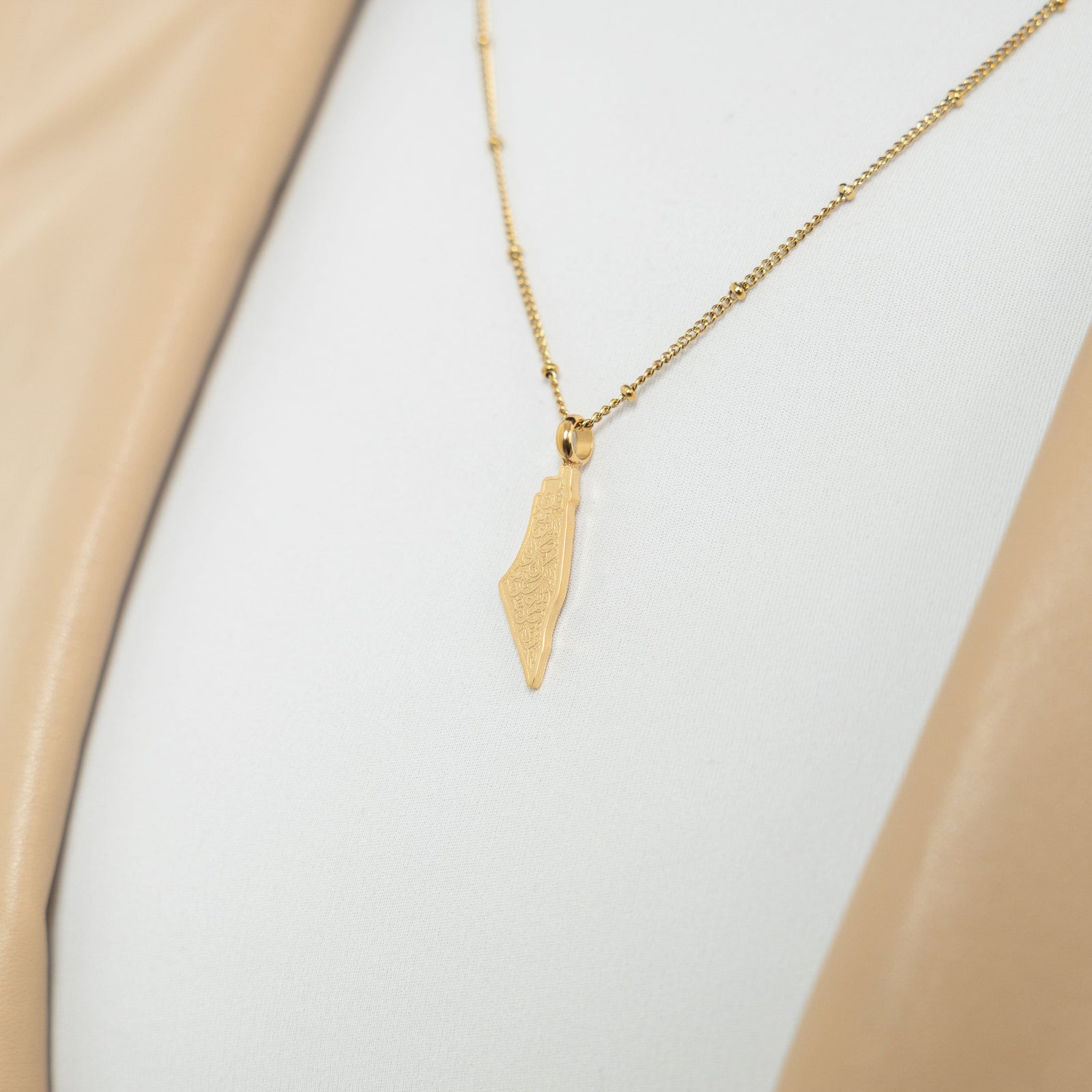 A model wearing the 18K gold-plated "With Hardship Comes Ease" Map Necklace. The necklace rests gracefully on her collarbone, highlighting the map pendant with Arabic calligraphy against a neutral-toned outfit.