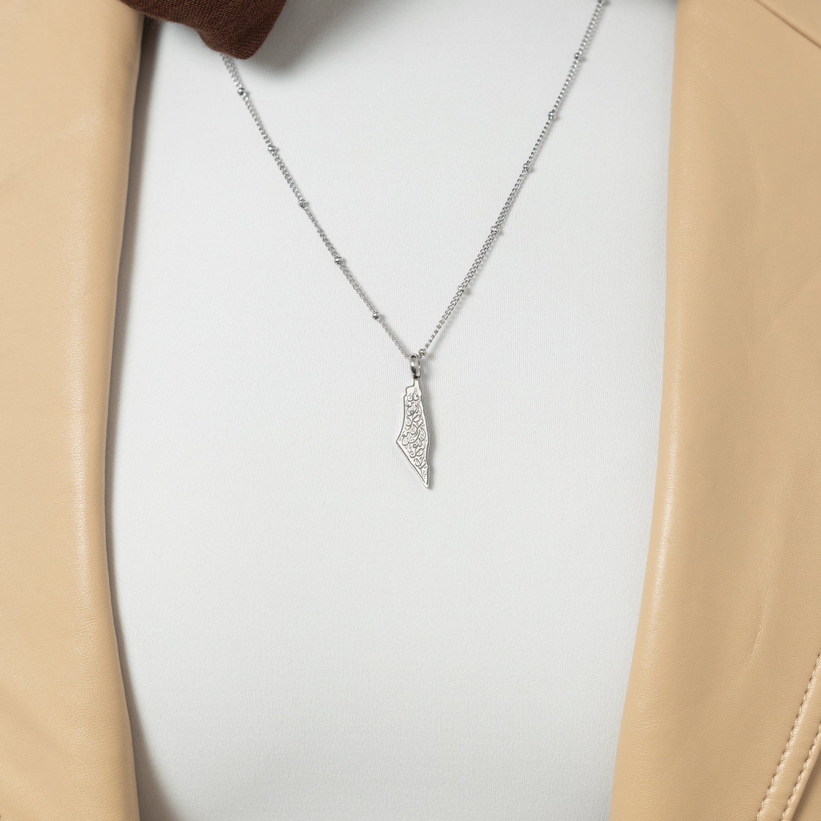 A model showcasing the silver "With Hardship Comes Ease" Map Necklace. The necklace sits delicately on her collarbone, emphasizing the map pendant with engraved Arabic script against a minimalist ensemble.