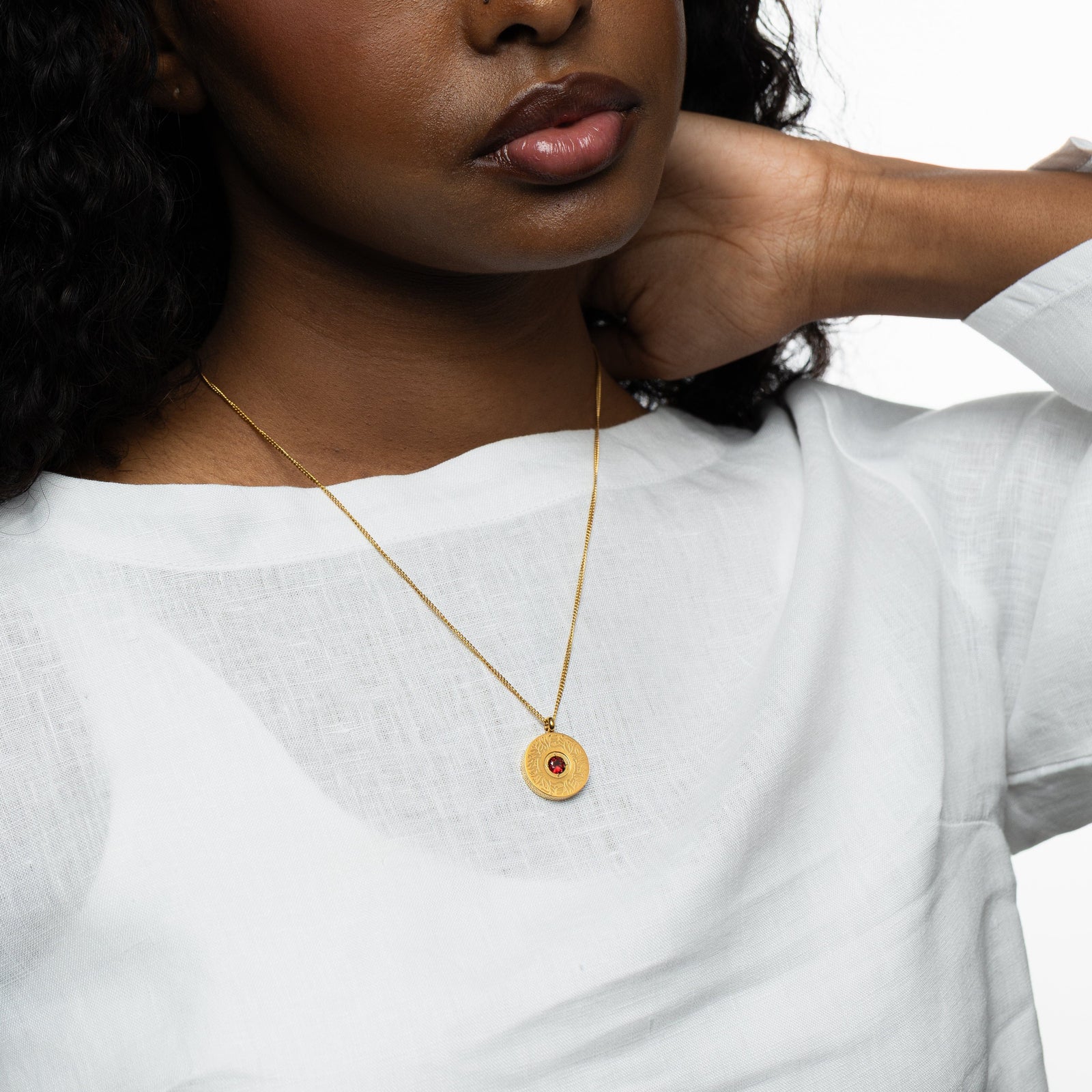 Mid-close view of the gold medallion necklace with red agate pendant resting over a white blouse, hand lifted near the neckline.