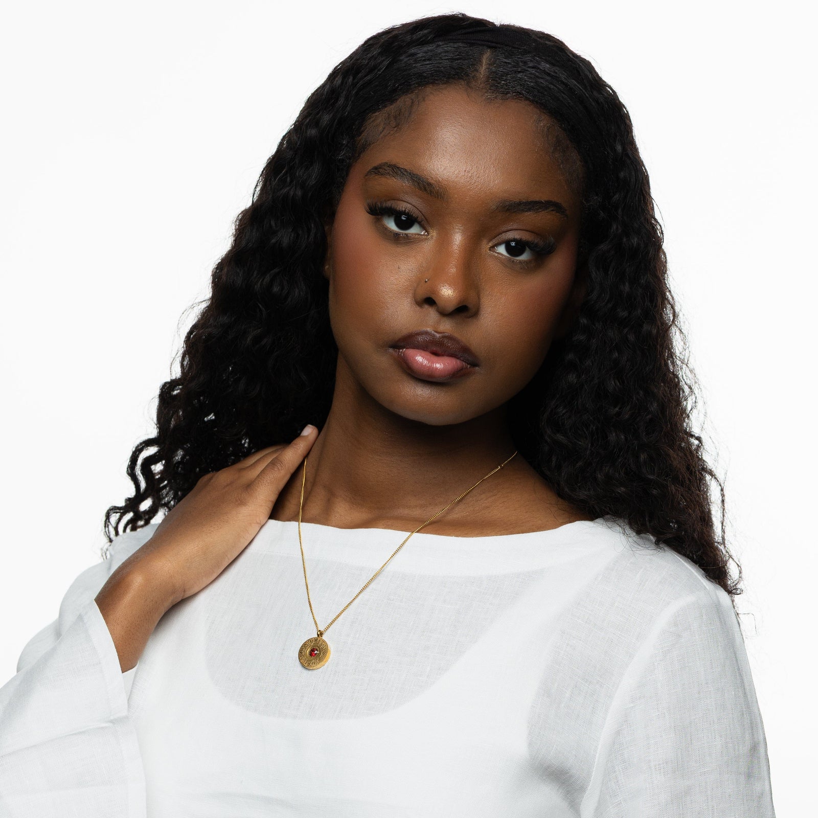 Woman wearing the gold medallion necklace with a red agate stone, styled with a white top, hand placed near her shoulder.