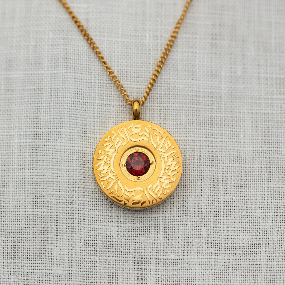 Close-up of the gold medallion pendant with a faceted red agate stone centerpiece and intricate Arabic calligraphy engraved in a circular design.