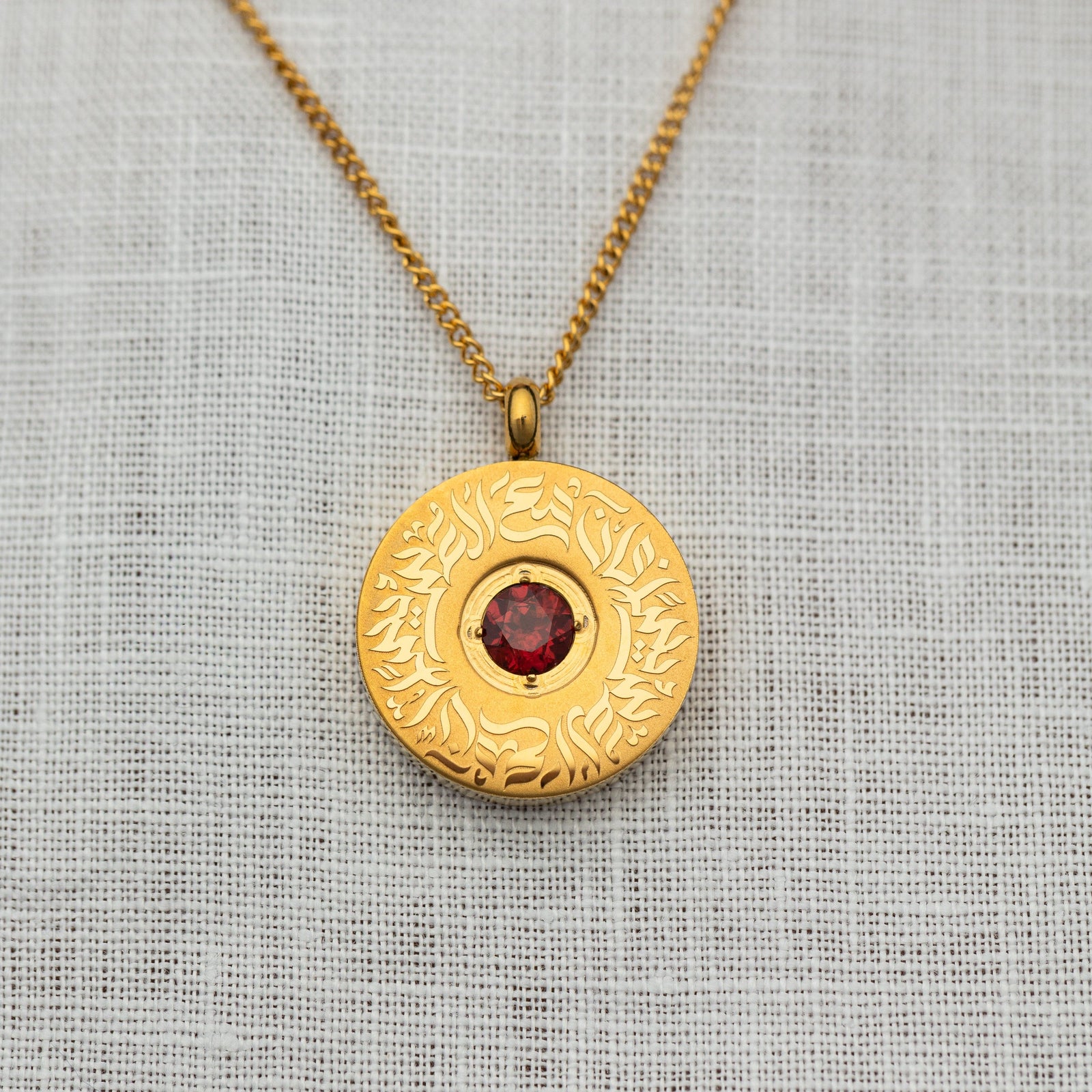 Close-up of the gold medallion pendant with a faceted red agate stone centerpiece and intricate Arabic calligraphy engraved in a circular design.