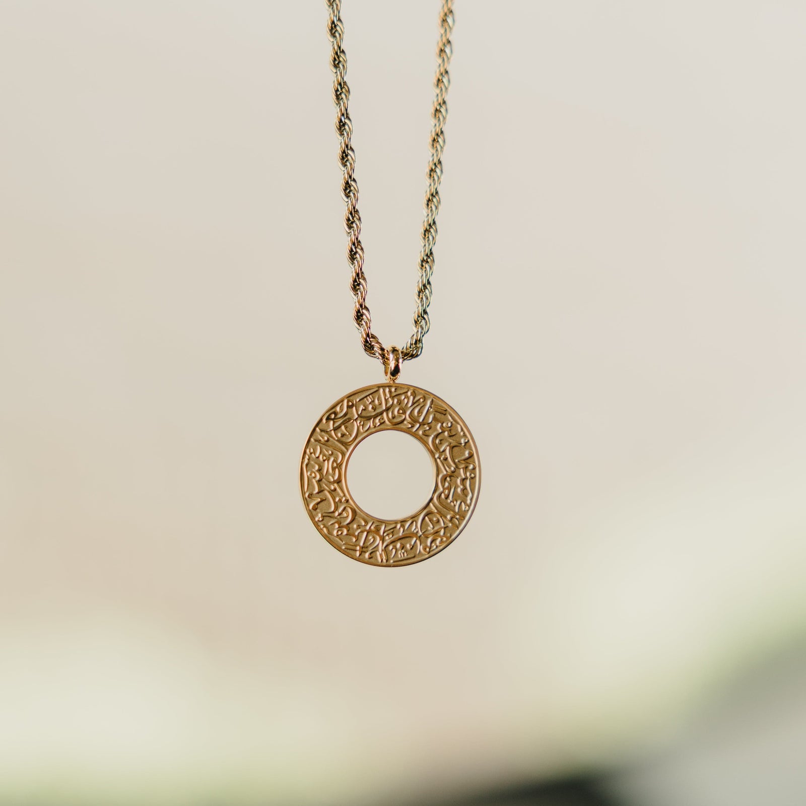 Gold car hanger with a circular pendant engraved in Arabic calligraphy reading “With Hardship Comes Ease,” suspended in focus against a soft background.