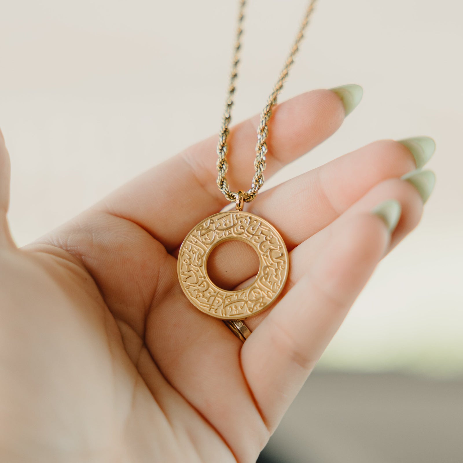 Hand holding a gold car hanger with a circular pendant engraved in Arabic calligraphy reading “With Hardship Comes Ease.”