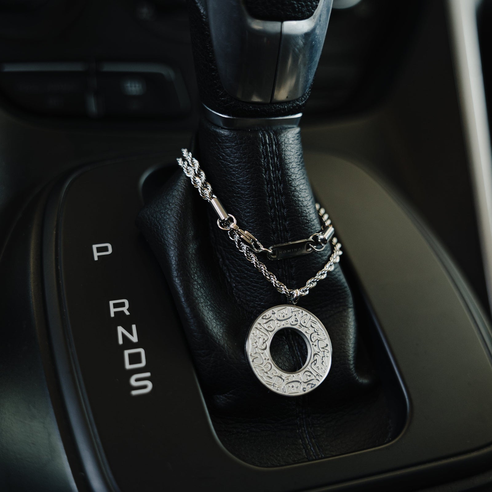 Silver car hanger with a circular pendant engraved in Arabic calligraphy reading “With Hardship Comes Ease,” draped around a black leather gear shift.