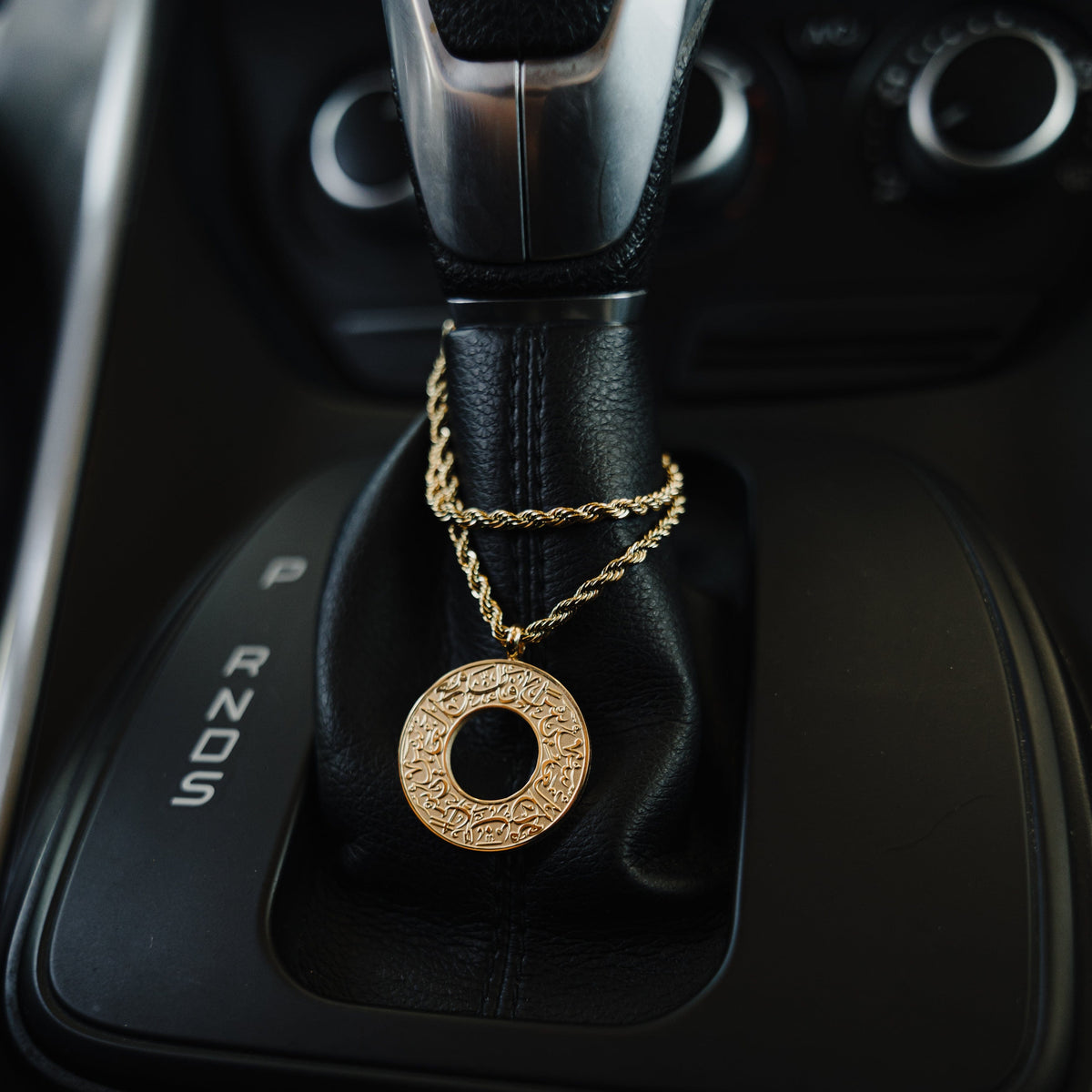 Gold car hanger with a circular pendant engraved in Arabic calligraphy reading “With Hardship Comes Ease,” draped around a black leather gear shift.