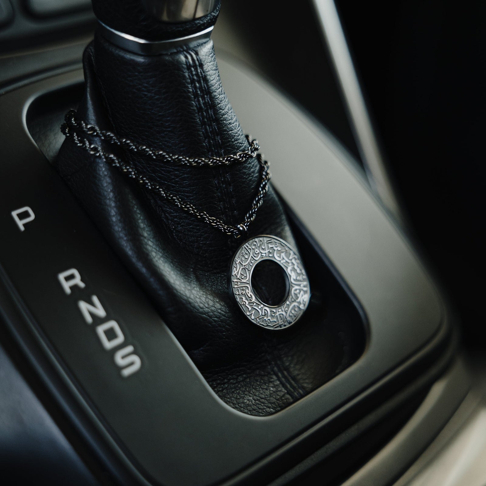 Black car hanger with a circular pendant engraved in Arabic calligraphy reading “With Hardship Comes Ease,” draped around a black leather gear shift.