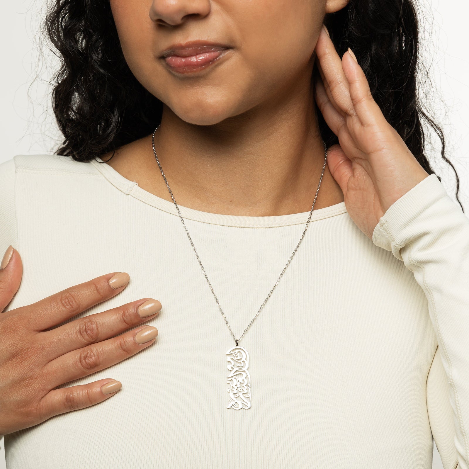 A model showcasing the silver "With Hardship Comes Ease" Calligraphy Necklace. The pendant hangs elegantly from her neck, emphasizing the detailed Arabic engraving against a neutral-toned outfit.