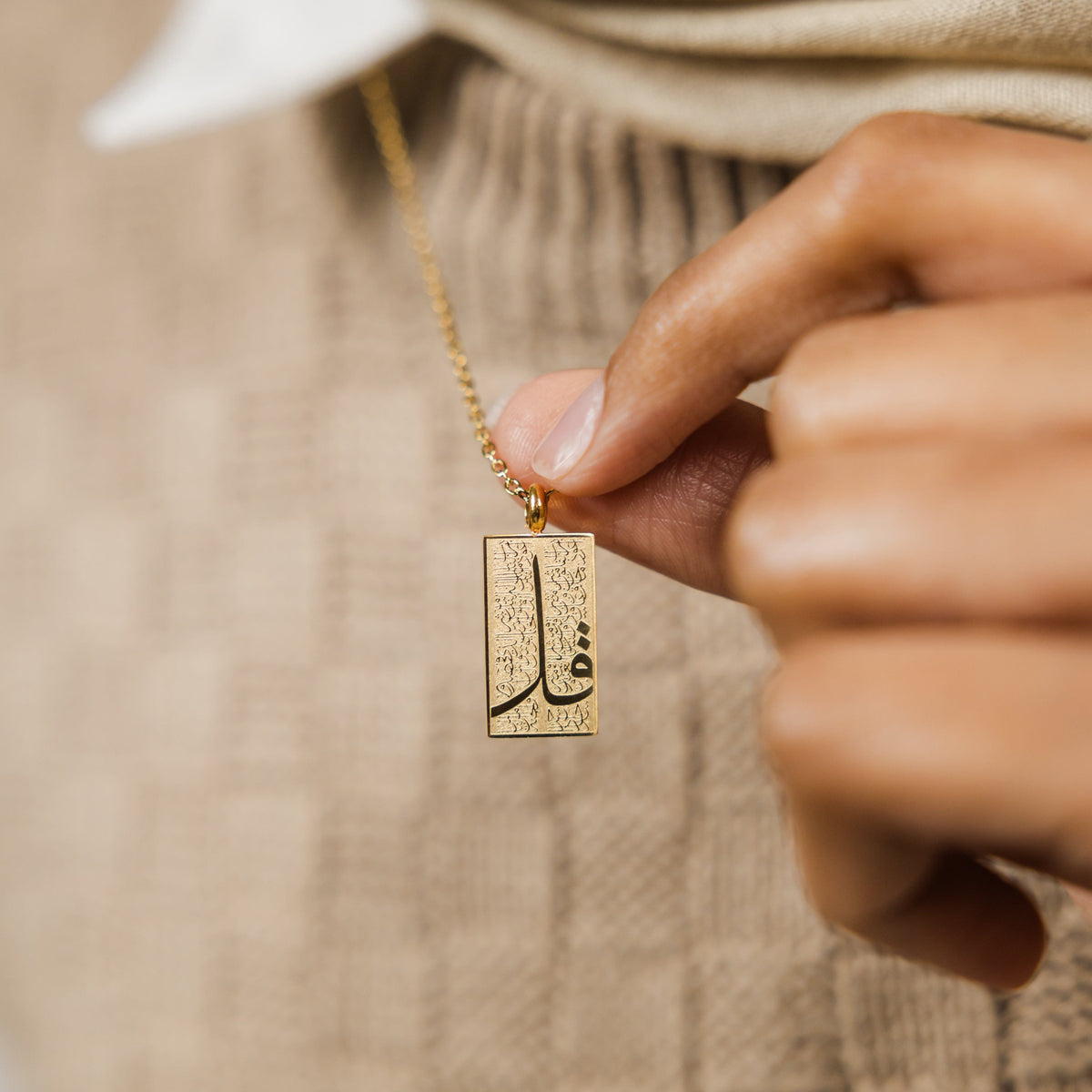 Close up of the gold '4 Qul' Tag Necklace as the model pulls the chain to better show the pendant.