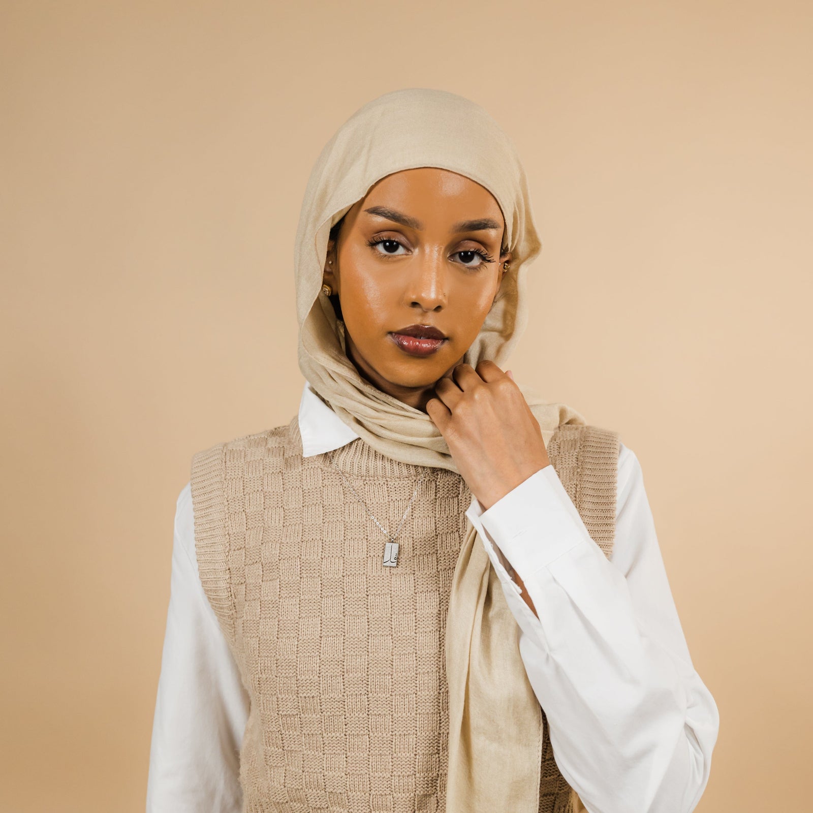 Photo of the model looking at the camera while holding their beige hijab with their left hand. The model is wearing the silver '4 Qul' Tag Necklace | Women.