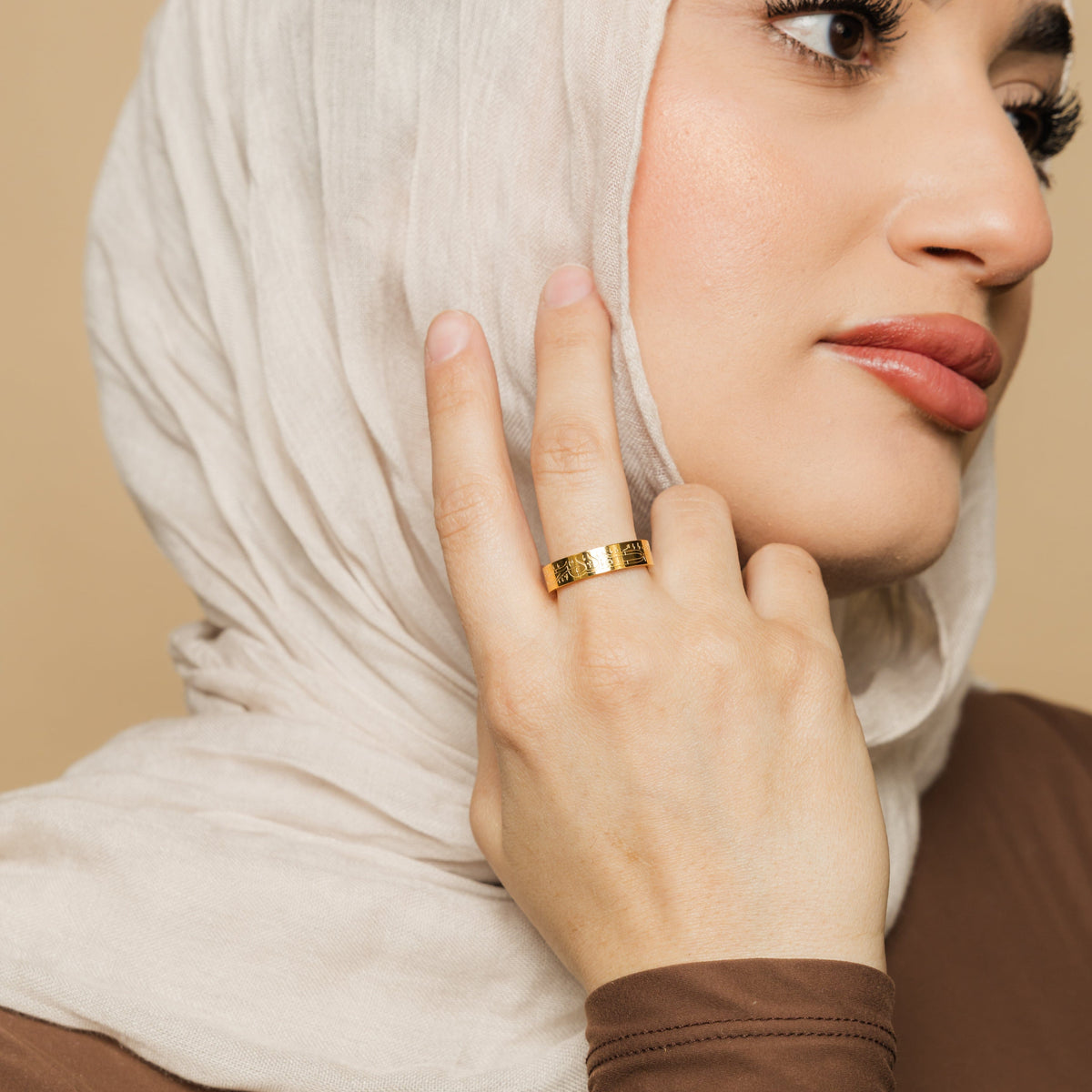 Woman wearing the gold Alhamdulillah ring on her middle finger, styled with a beige hijab and brown top, hand resting on her face.