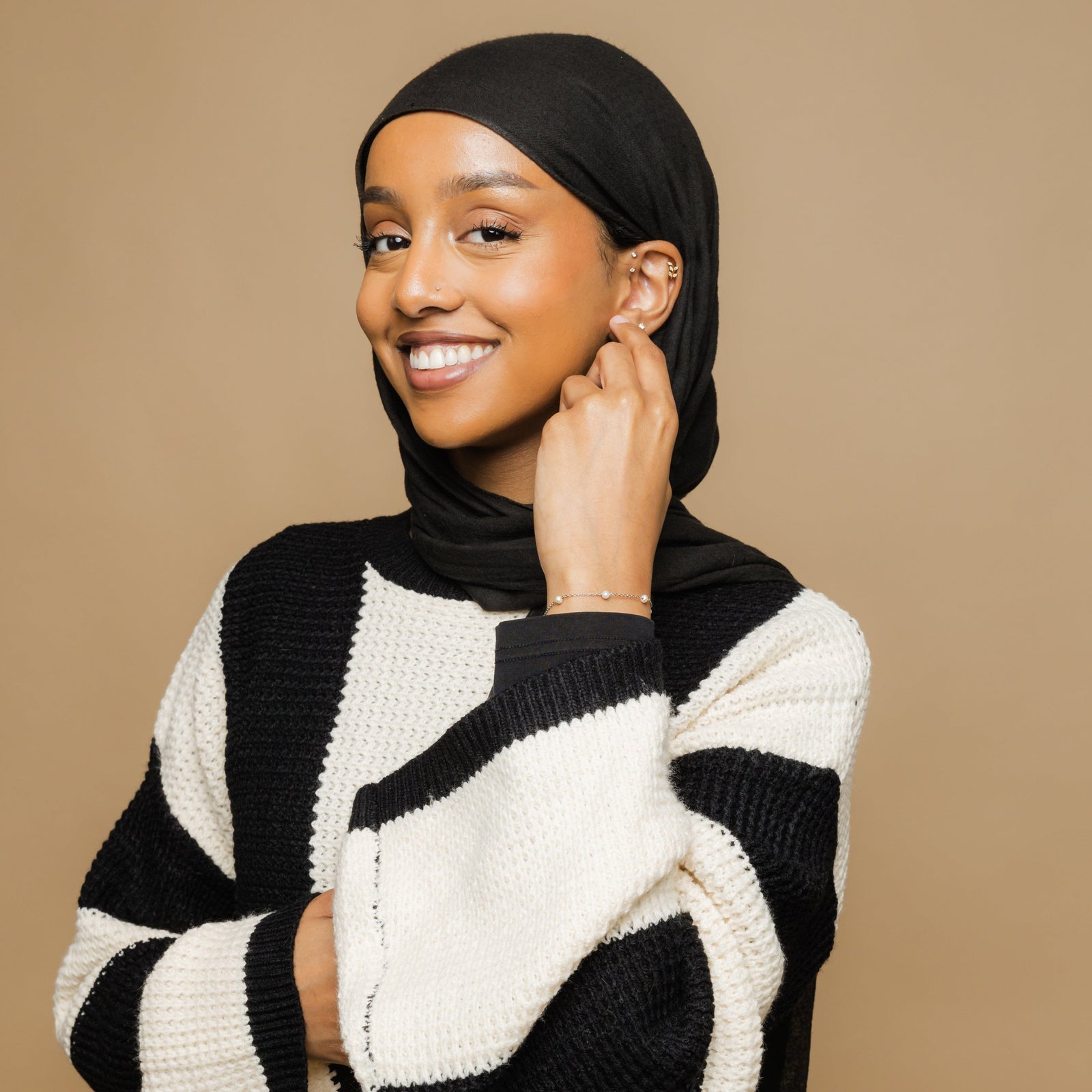 Photo of the model smiling to the camera while touching their left ear with their left hand. The model is wearing the Pearl Strand Bracelet on their left wrist.