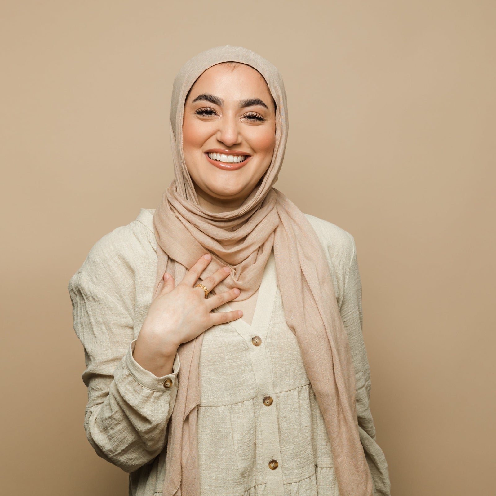 Woman in a beige hijab and buttoned tunic, hand on chest showcasing the gold Five Pillars Ring.