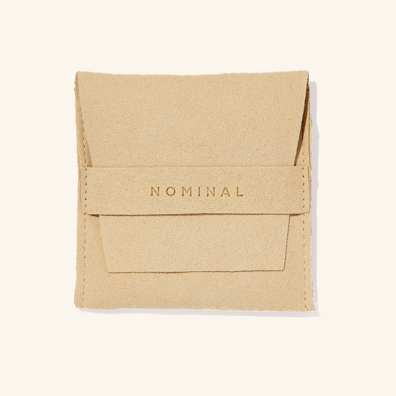 The image shows a small beige suede pouch placed against a light, neutral background. The pouch has a soft, textured surface and a flap closure at the top.

Centered on the front flap, the word “NOMINAL” is embossed in capital letters, giving it a subtle, refined look. The pouch appears neatly stitched along the edges and has a clean, minimalist design, suggesting it’s used for packaging or storing jewelry or small accessories.
