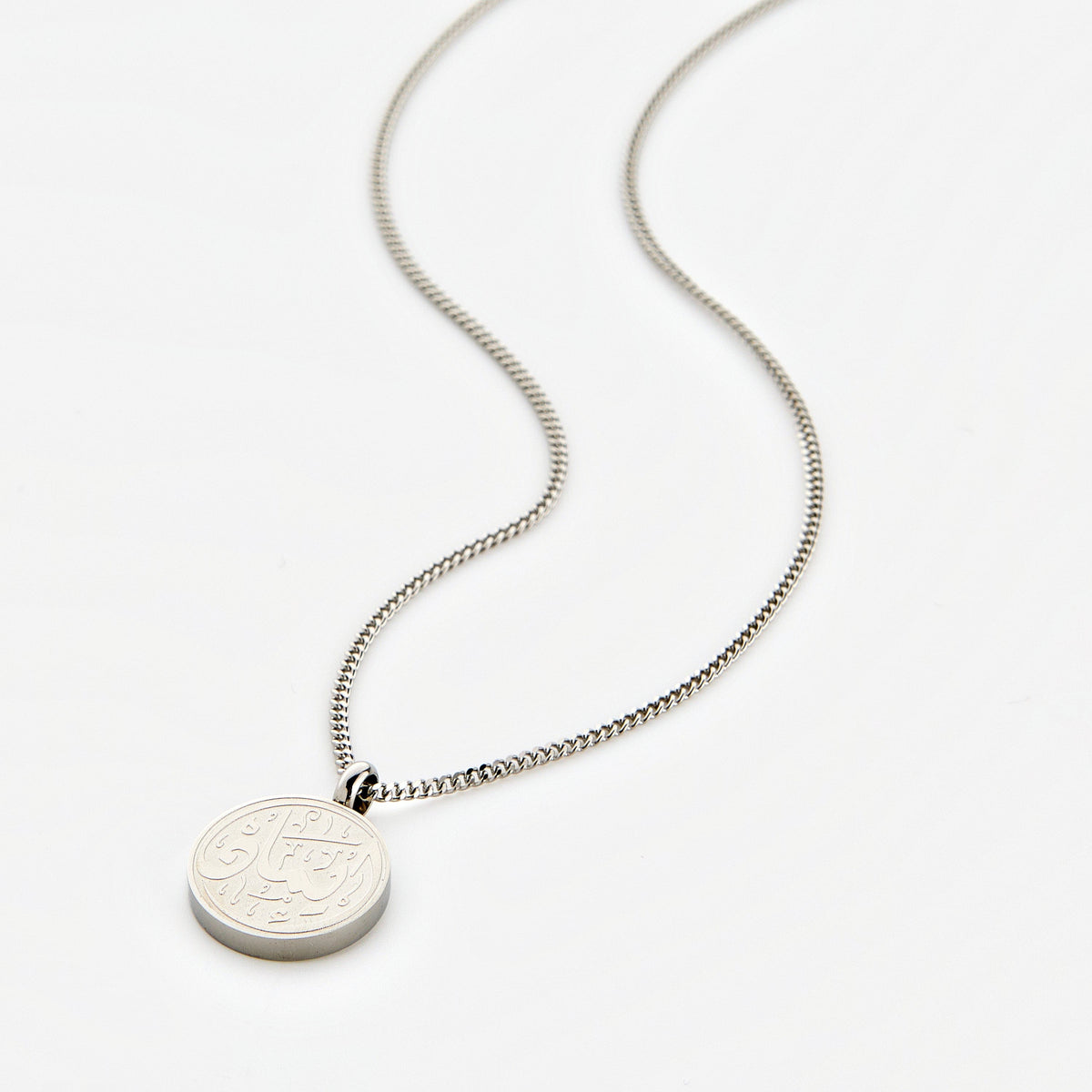A silver Faith Token necklace with the pendant resting flat on a light background, showing the engraved Arabic script “Iman”.