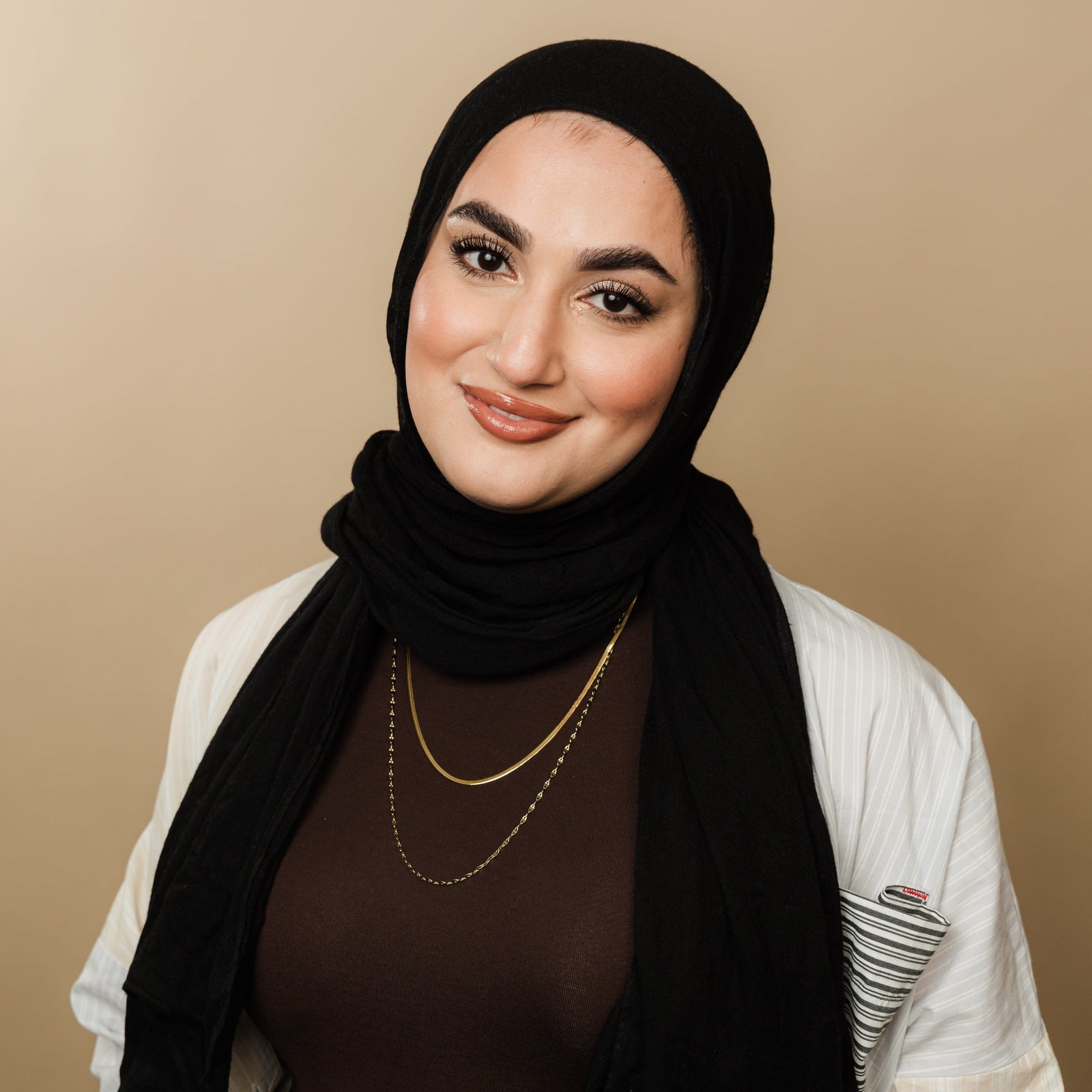 Woman wearing a brown top, black hijab, striped detail shirt, and layered gold necklaces with a herringbone chain and flutter chain.