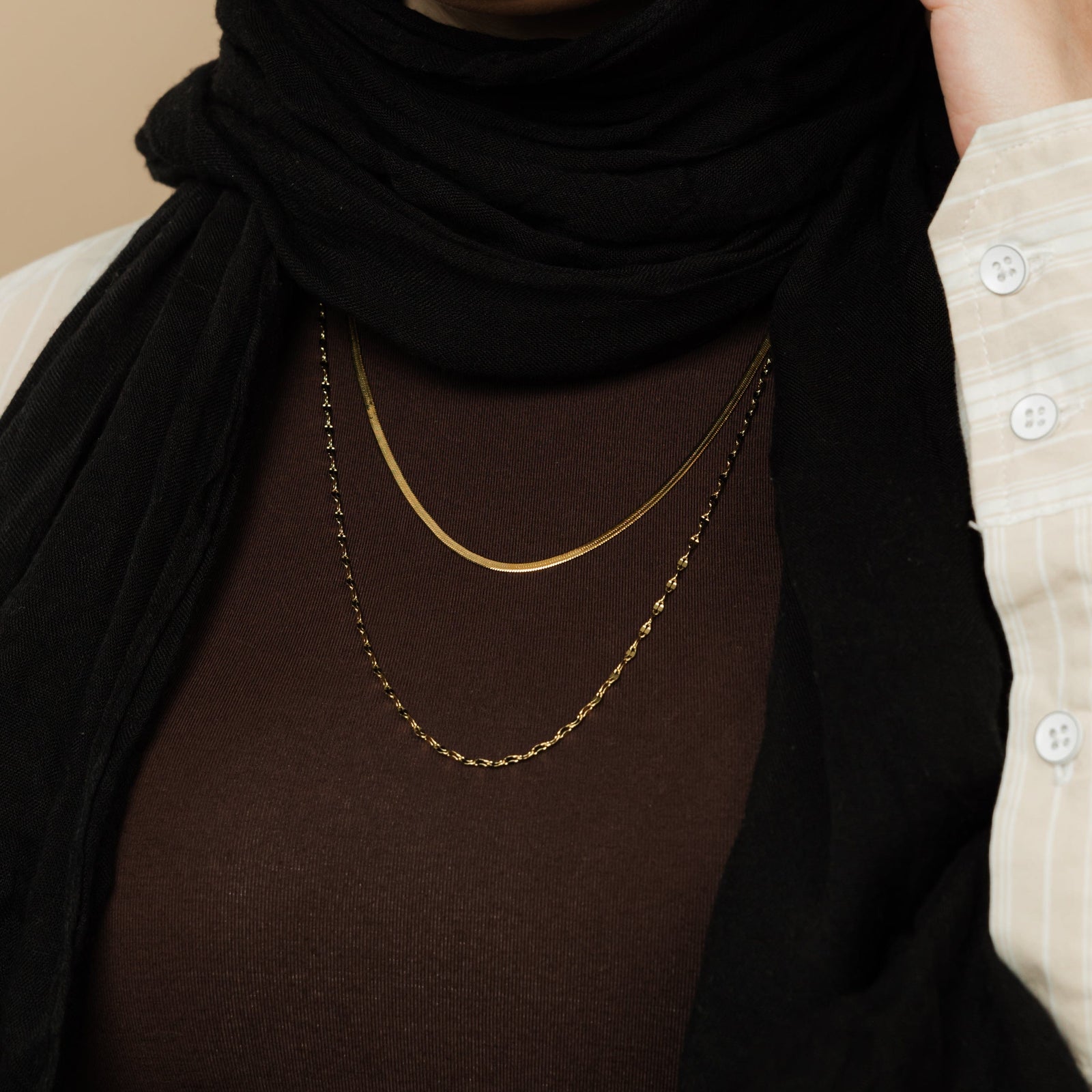 Close-up of a gold herringbone chain and flutter chain layered over a brown top, worn by a woman styled with a black hijab and striped detail shirt.