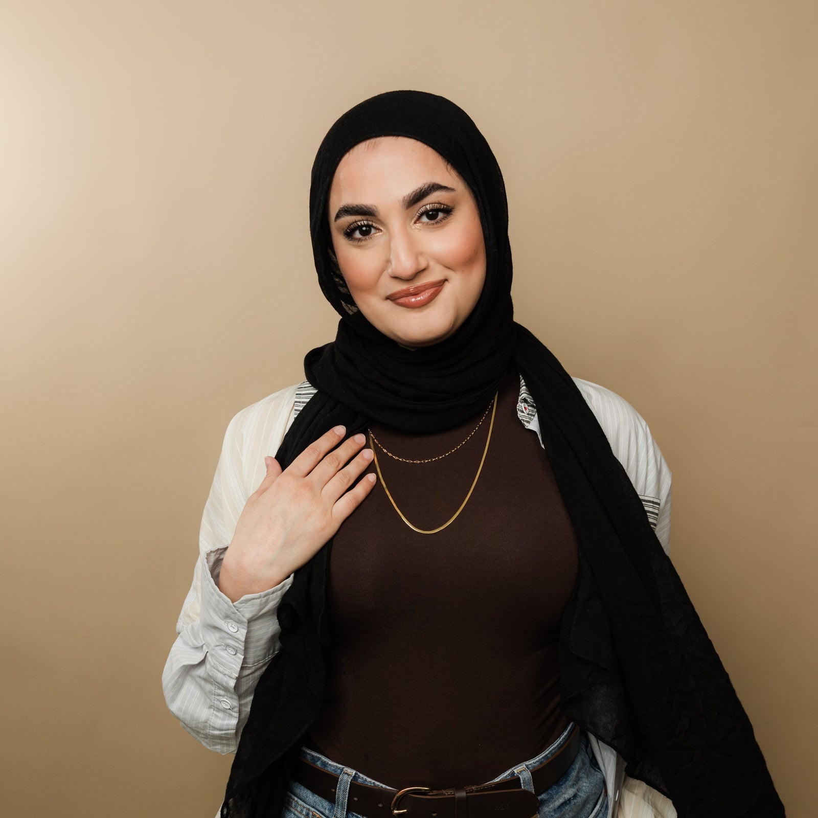 Woman wearing a brown top, black hijab, striped detail shirt, and layered gold necklaces featuring a herringbone chain and flutter chain.