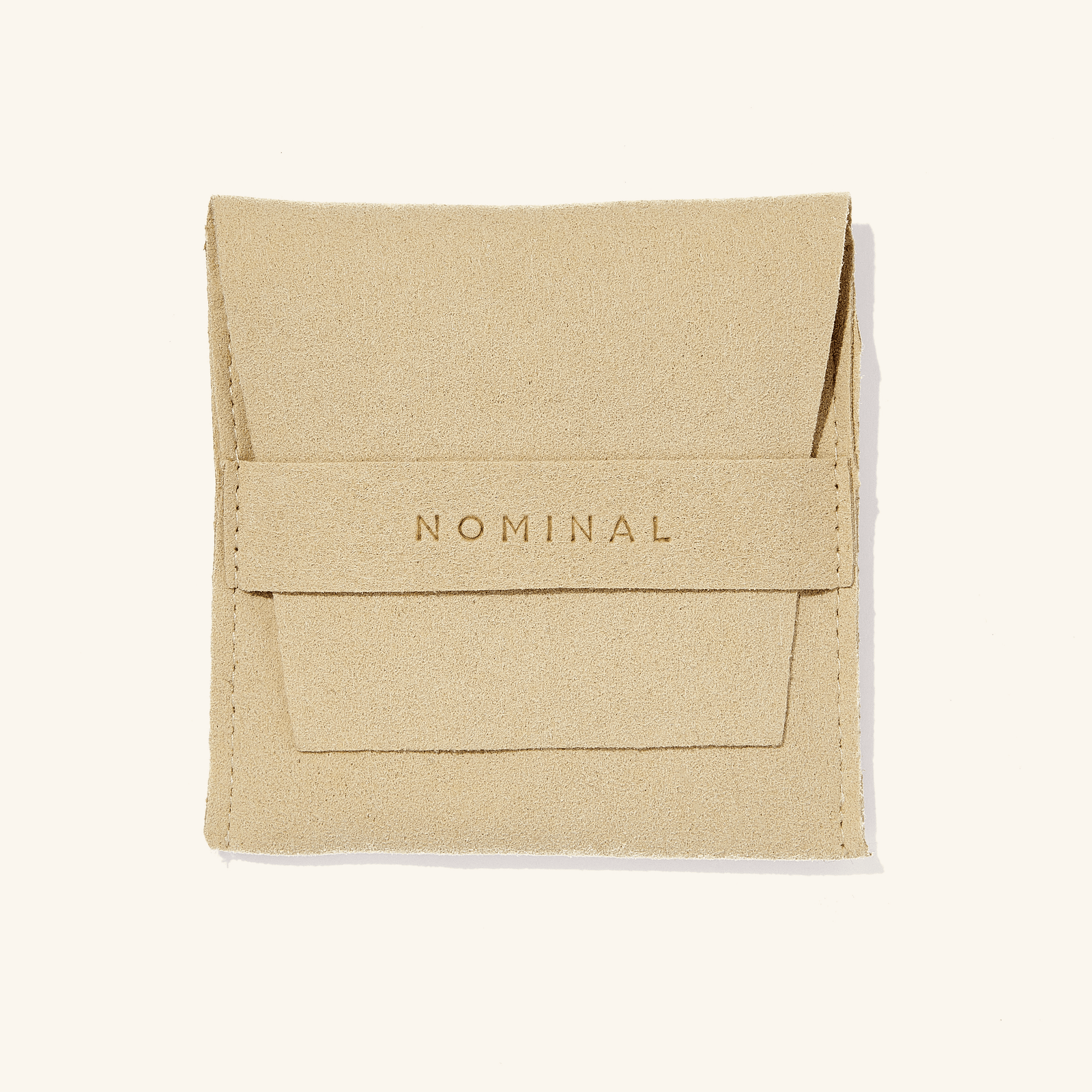 The image shows a small beige suede pouch placed against a light, neutral background. The pouch has a soft, textured surface and a flap closure at the top.

Centered on the front flap, the word “NOMINAL” is embossed in capital letters, giving it a subtle, refined look. The pouch appears neatly stitched along the edges and has a clean, minimalist design, suggesting it’s used for packaging or storing jewelry or small accessories.